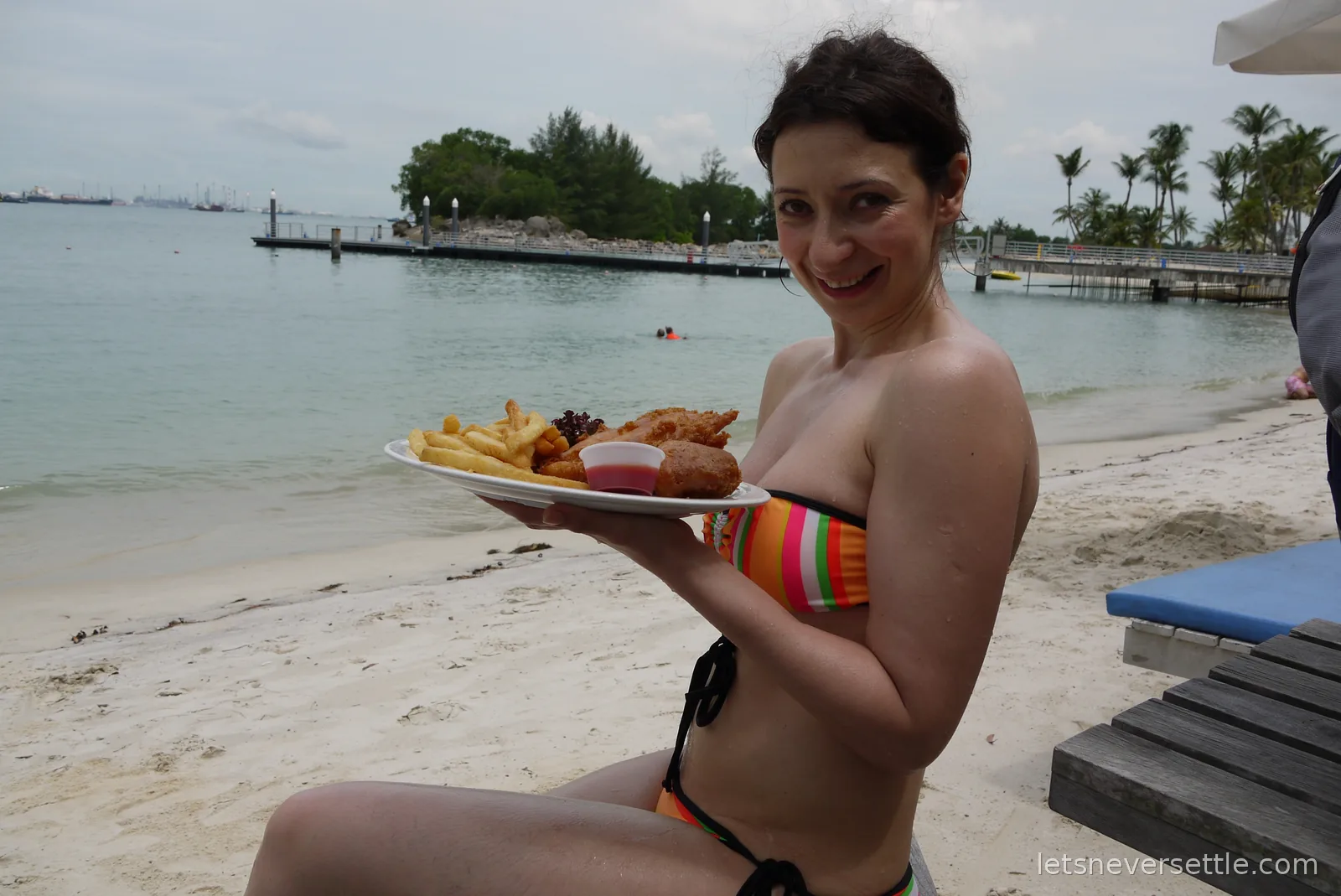 Inga holding a plate of fish and chips on the sand at Siloso Beach, with the swimming lagoon and pier behind
