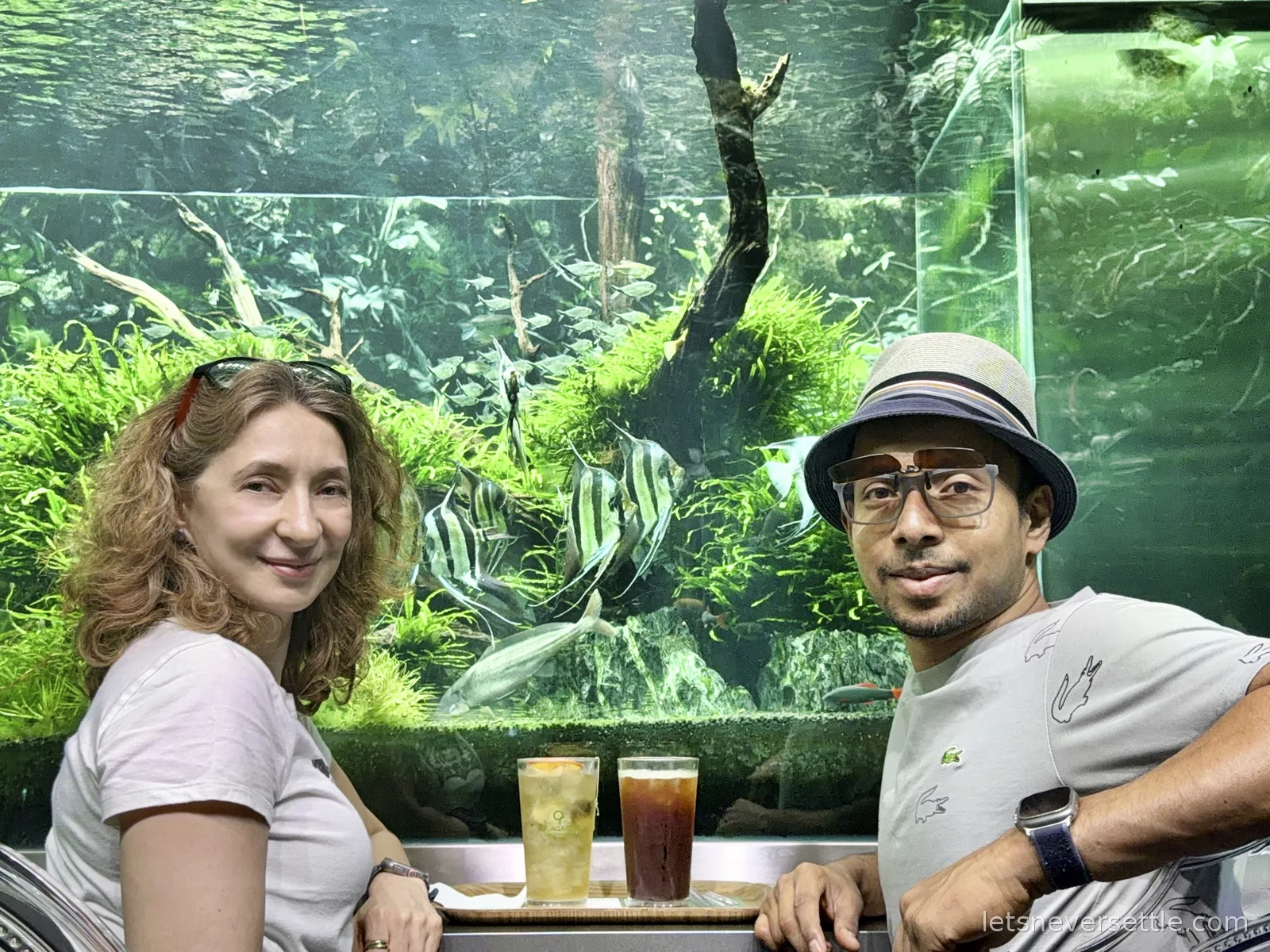 Inga and Dileep sitting with two iced drinks at a counter built against a large planted aquarium full of angelfish