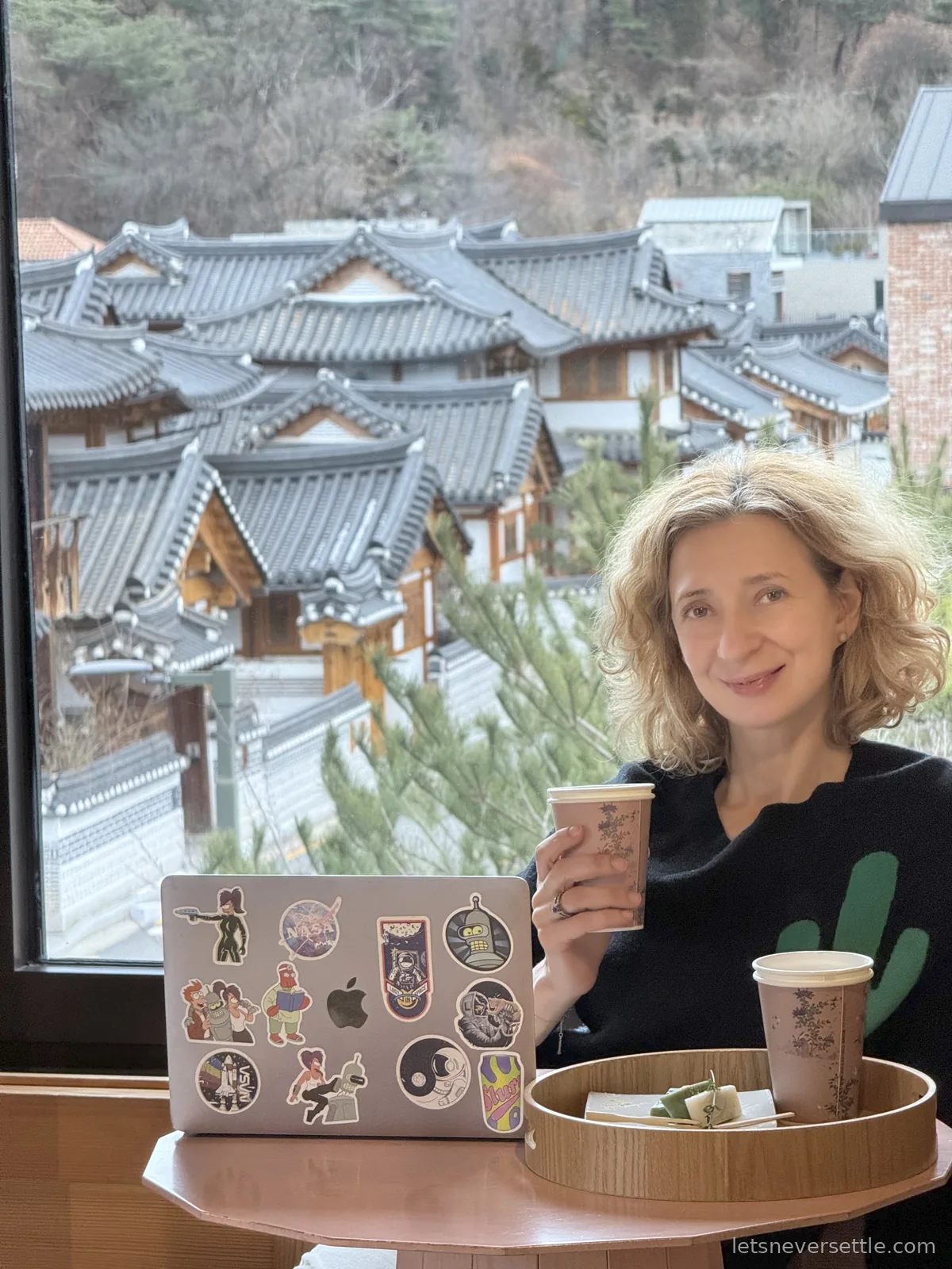 Inga with a coffee and a sticker-covered laptop at a cafe window overlooking hanok rooftops and pine trees