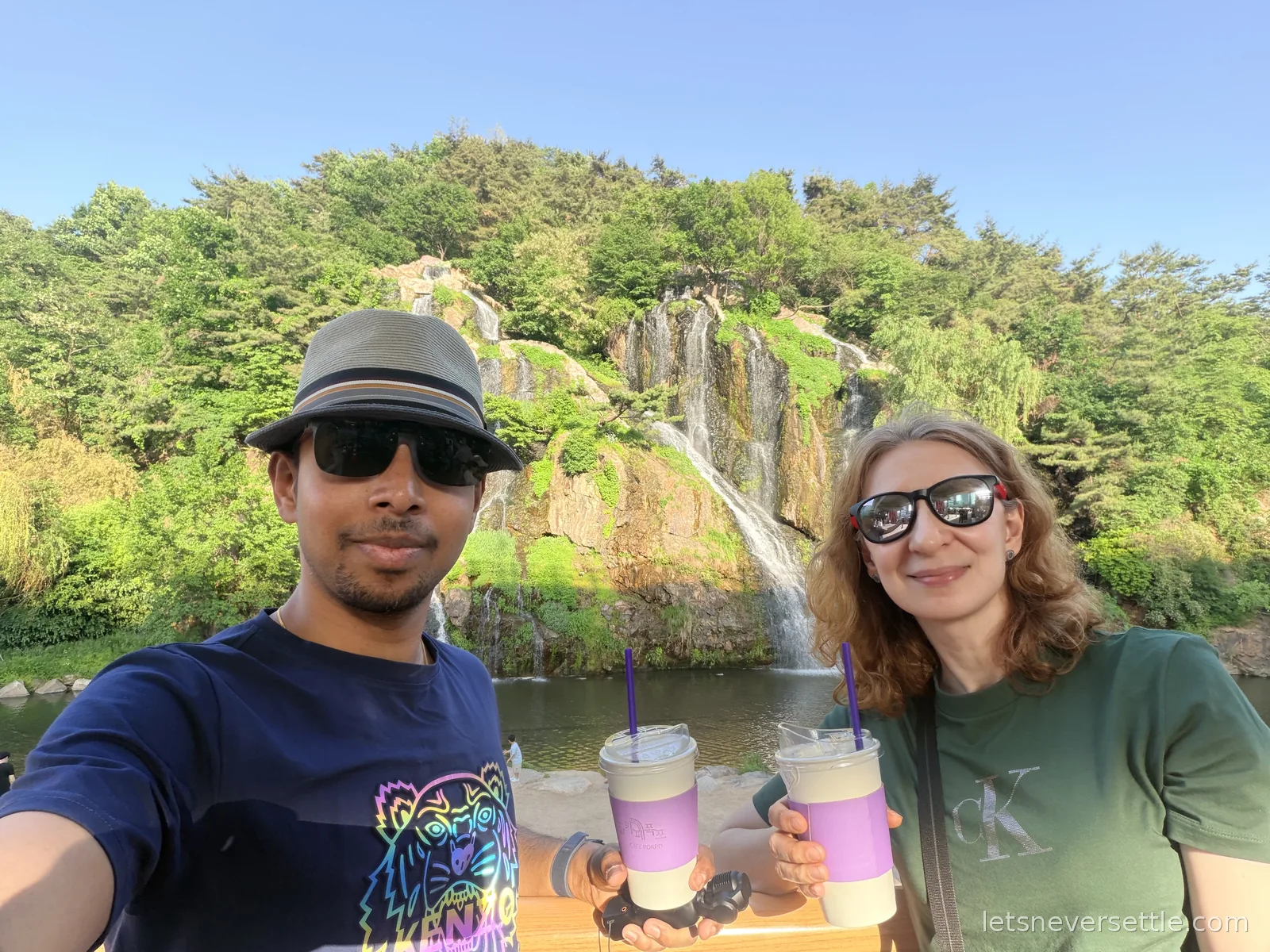 Selfie of Inga and Dileep holding iced drinks in front of the Hongjecheon artificial waterfall