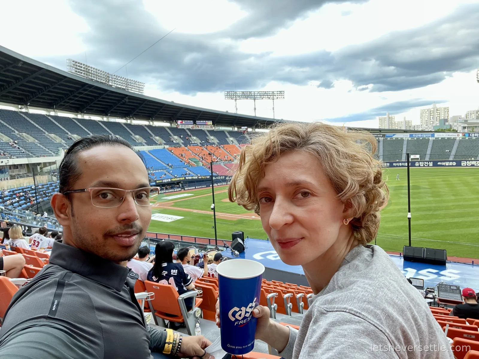 Selfie of Inga and Dileep in the stands at Jamsil Baseball Stadium, holding a blue Cass beer cup