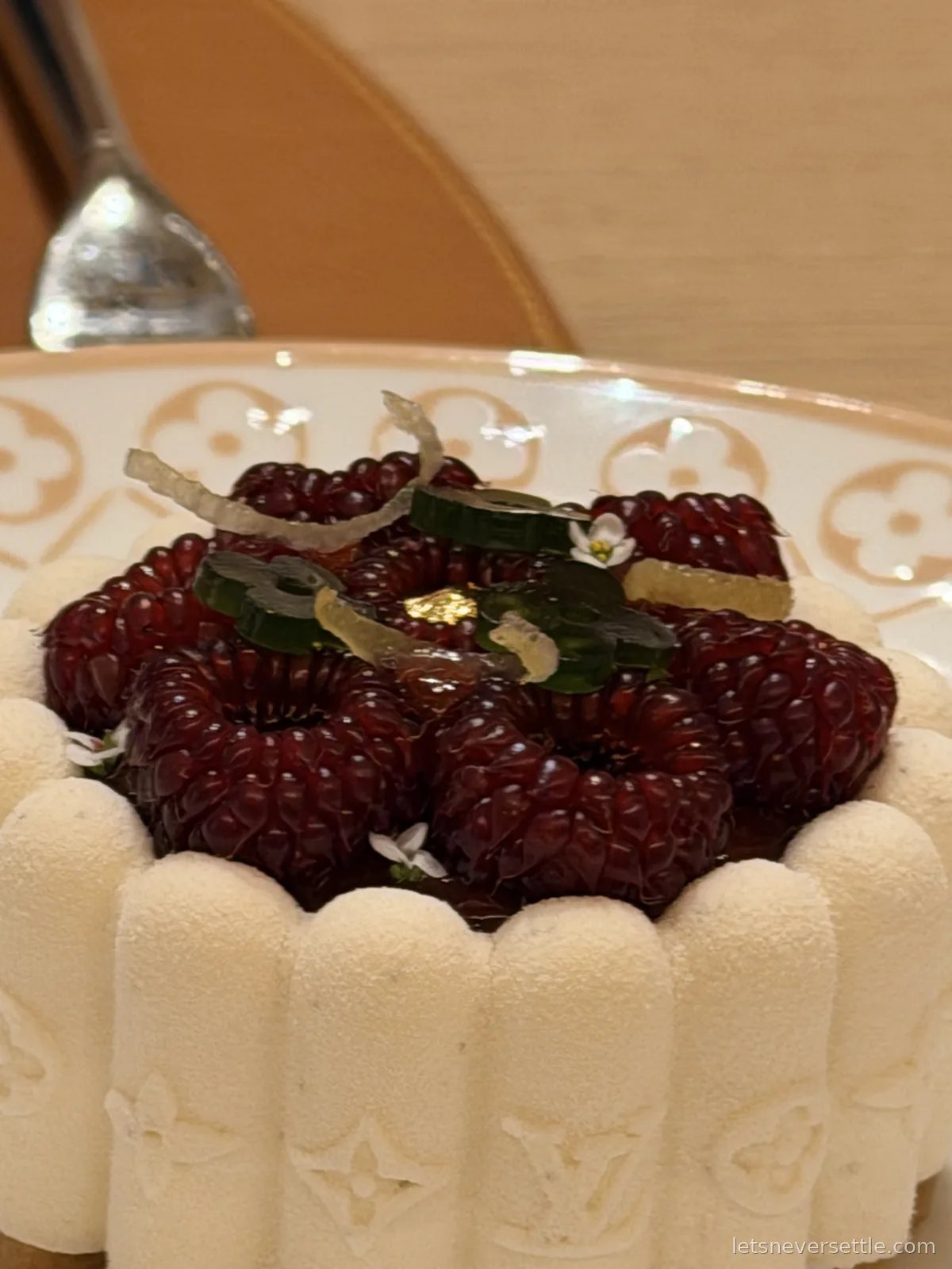 A pale mousse cake topped with dark red berries and candied peel, its sides shaped into monogrammed petals