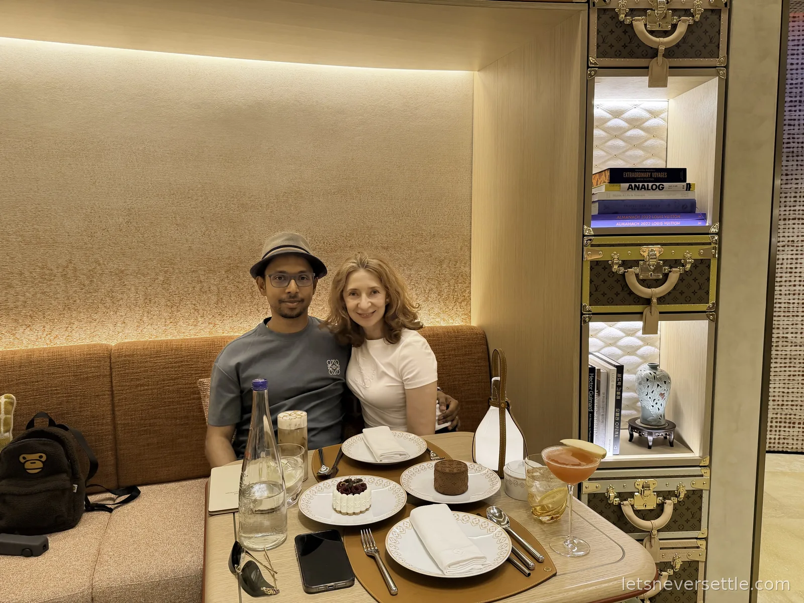 Inga and Dileep sitting together in a curved booth at the Louis Vuitton cafe in Seoul, a display of monogrammed trunks and books beside them