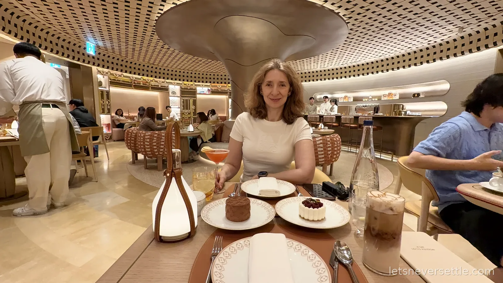 Inga at a table of desserts in the Louis Vuitton cafe in Seoul, a sculptural bronze column and woven ceiling above the dining room