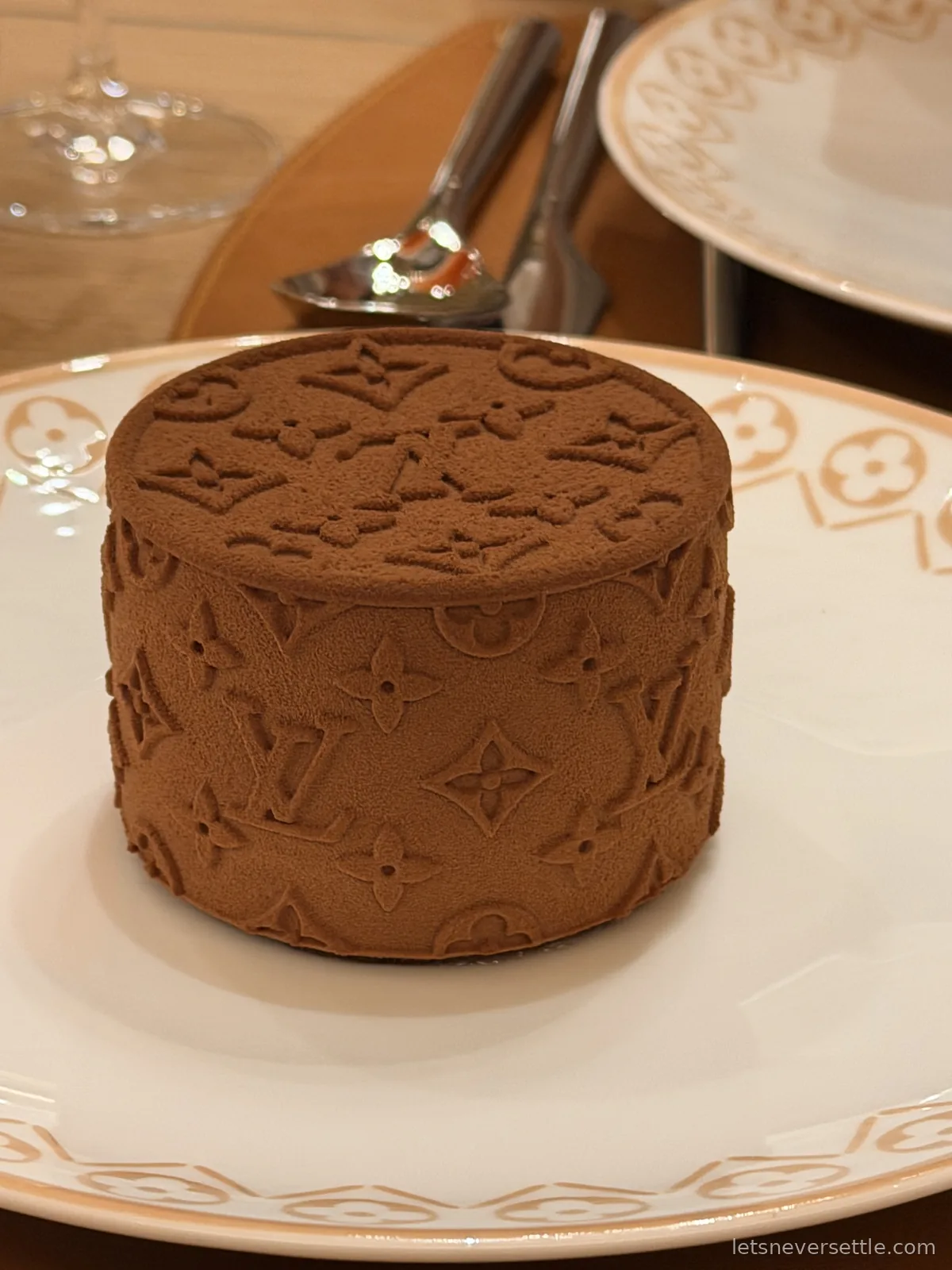 A small round chocolate cake covered in cocoa and embossed with the Louis Vuitton monogram, on a matching monogrammed plate