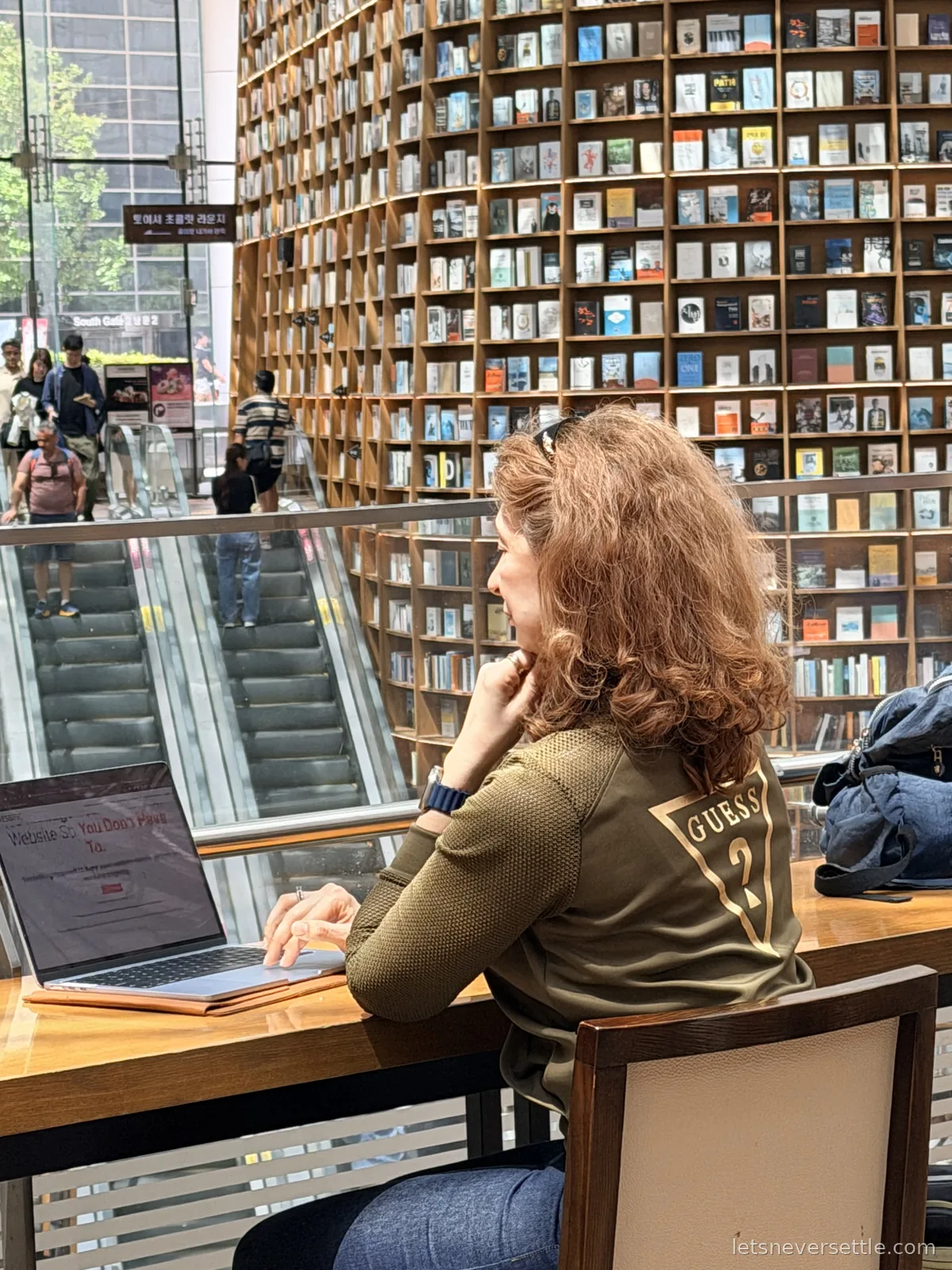 Inga working at a long wooden counter in Starfield Library, the towering wall of books and the mall escalators behind her