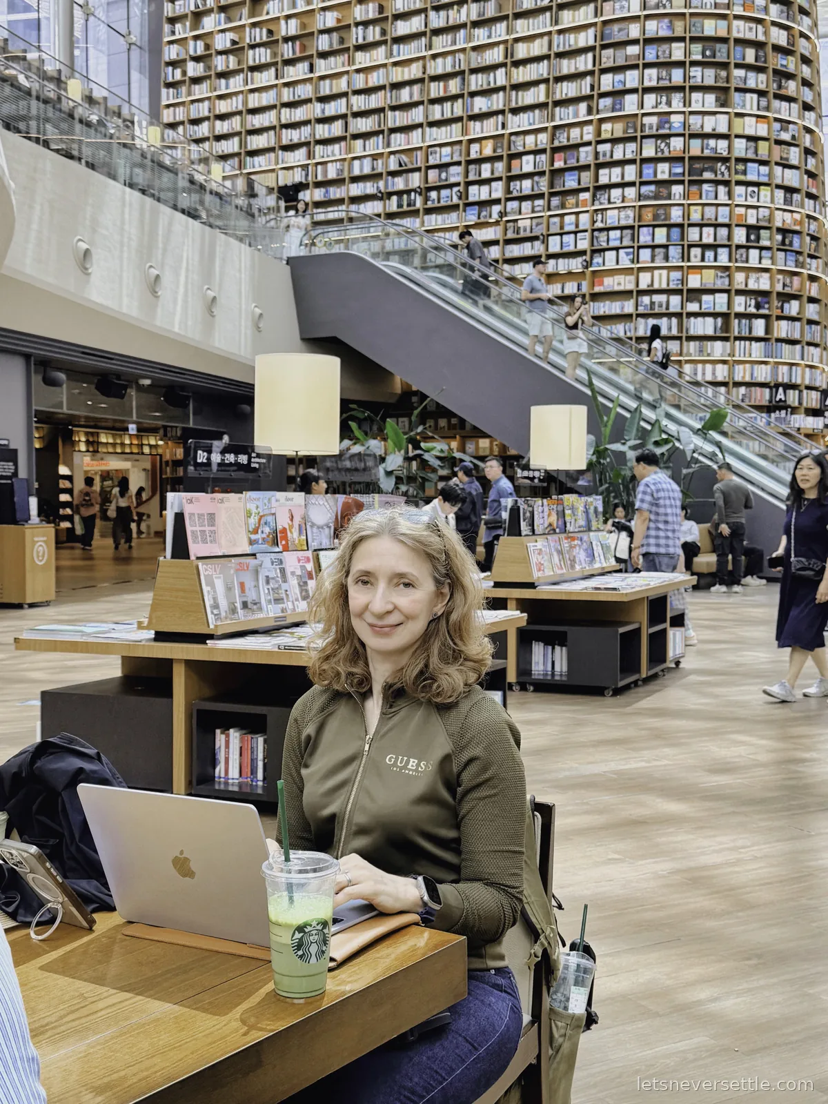 Inga working on a laptop with an iced green drink at a desk in Starfield Library, bookshelves rising behind