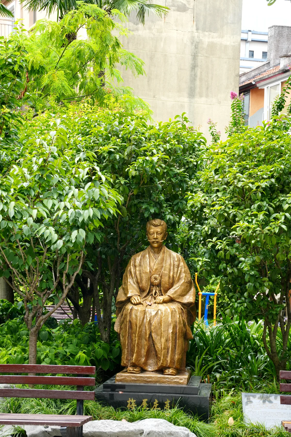 A seated bronze statue of Umeya Shokichi in a kimono among dense greenery, his name in characters on the black stone base