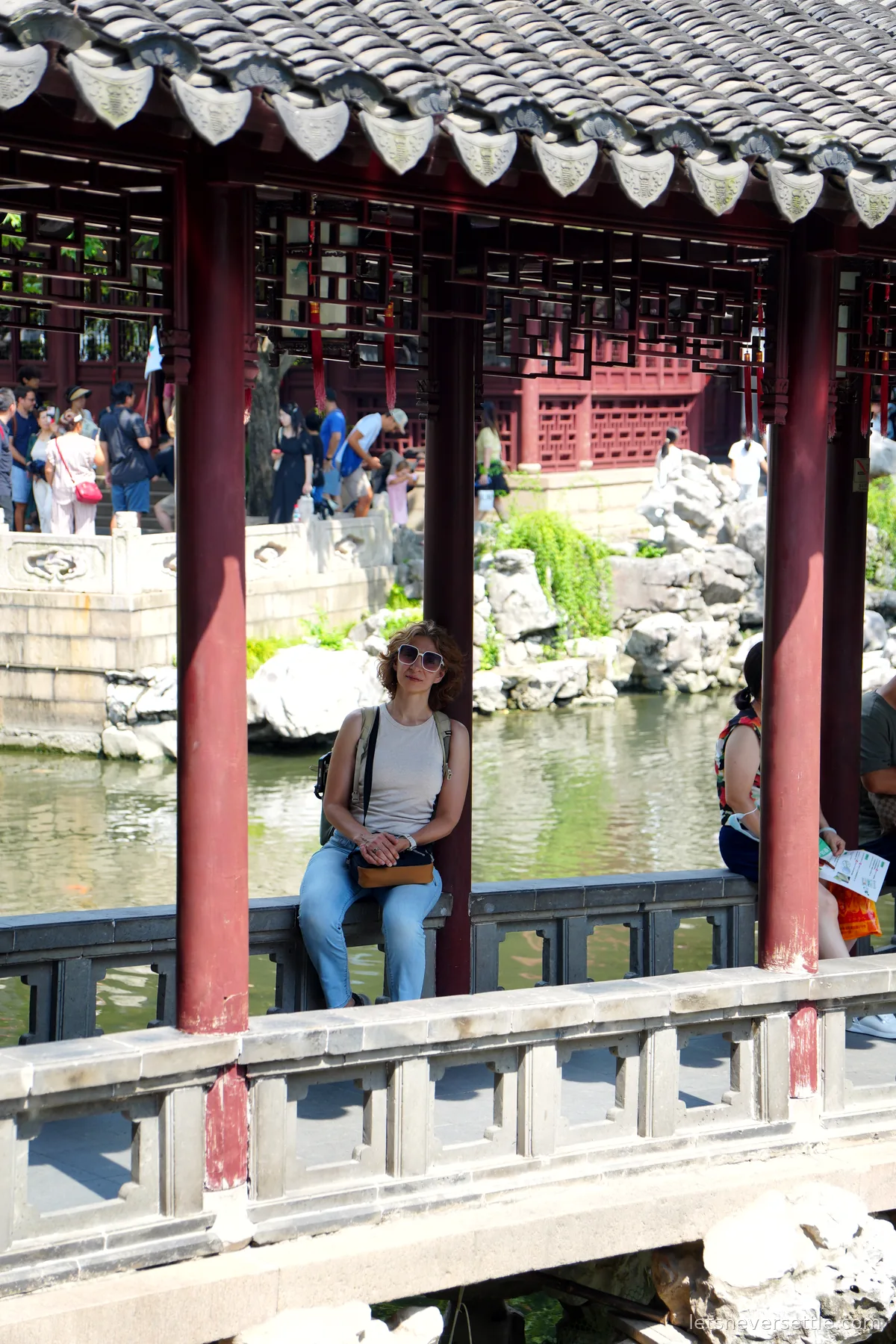 Inga sitting on the stone balustrade of a covered corridor in Yu Garden, Taihu rocks and a green pond behind her