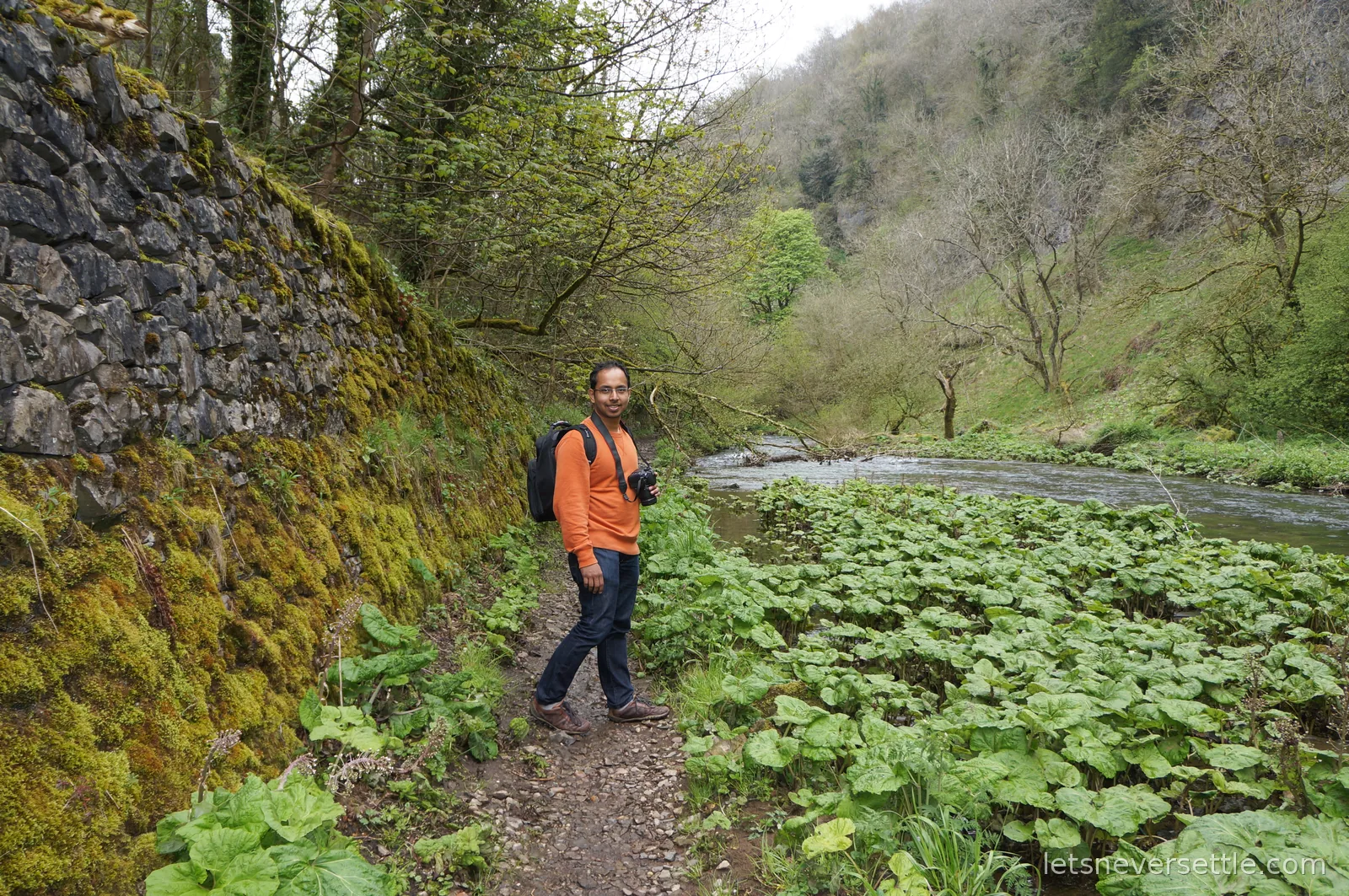 Dileep with a backpack and camera on a stony riverside path in a Peak District dale, a moss-covered stone wall rising beside him