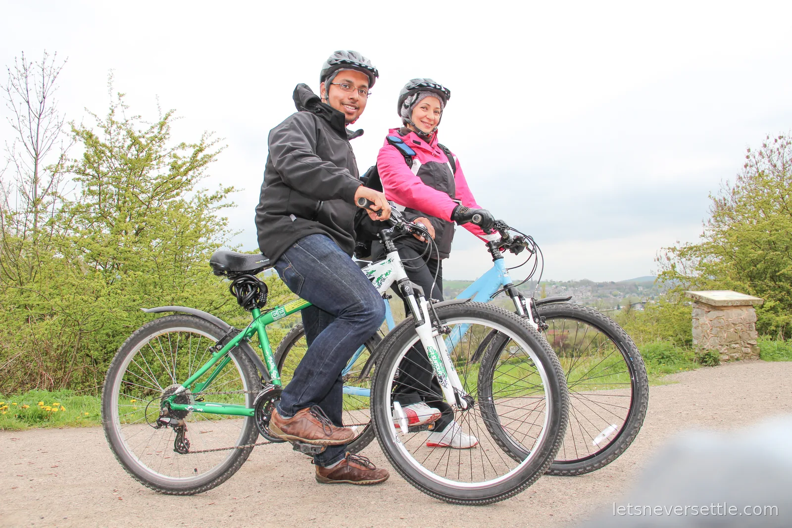 Dileep and Inga wearing helmets with two hired mountain bikes on the Monsal Trail, a green valley and town below behind them