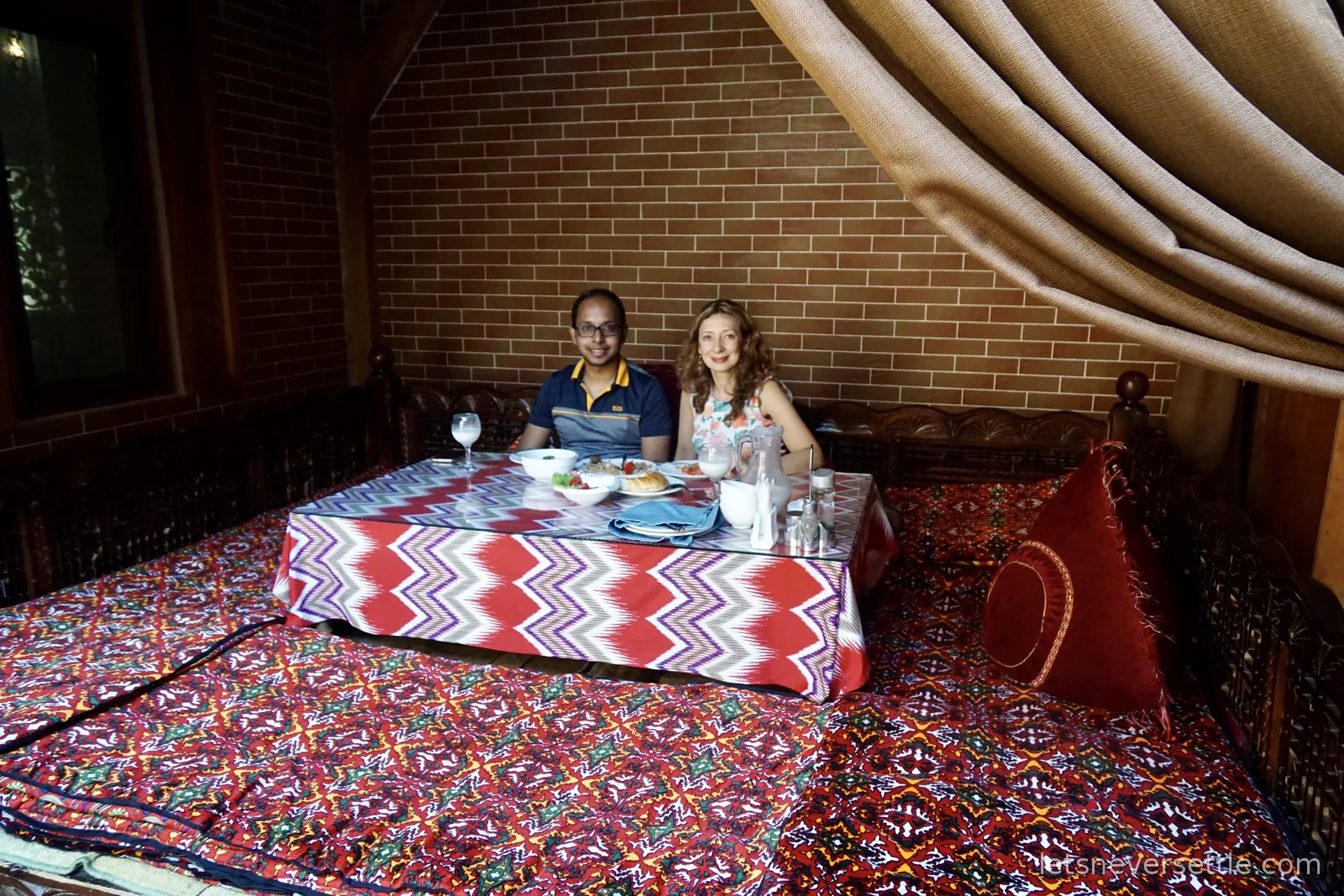 Inga and Dileep at a low glass-topped table with a zigzag ikat cloth, set on carpeted daybed seating with a jug of milky drink and dishes