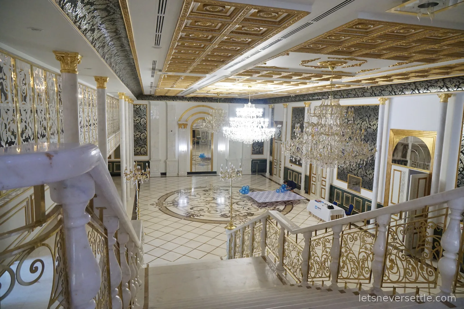A marble staircase with gilded railings descending into a hotel hall with a gold coffered ceiling and three crystal chandeliers
