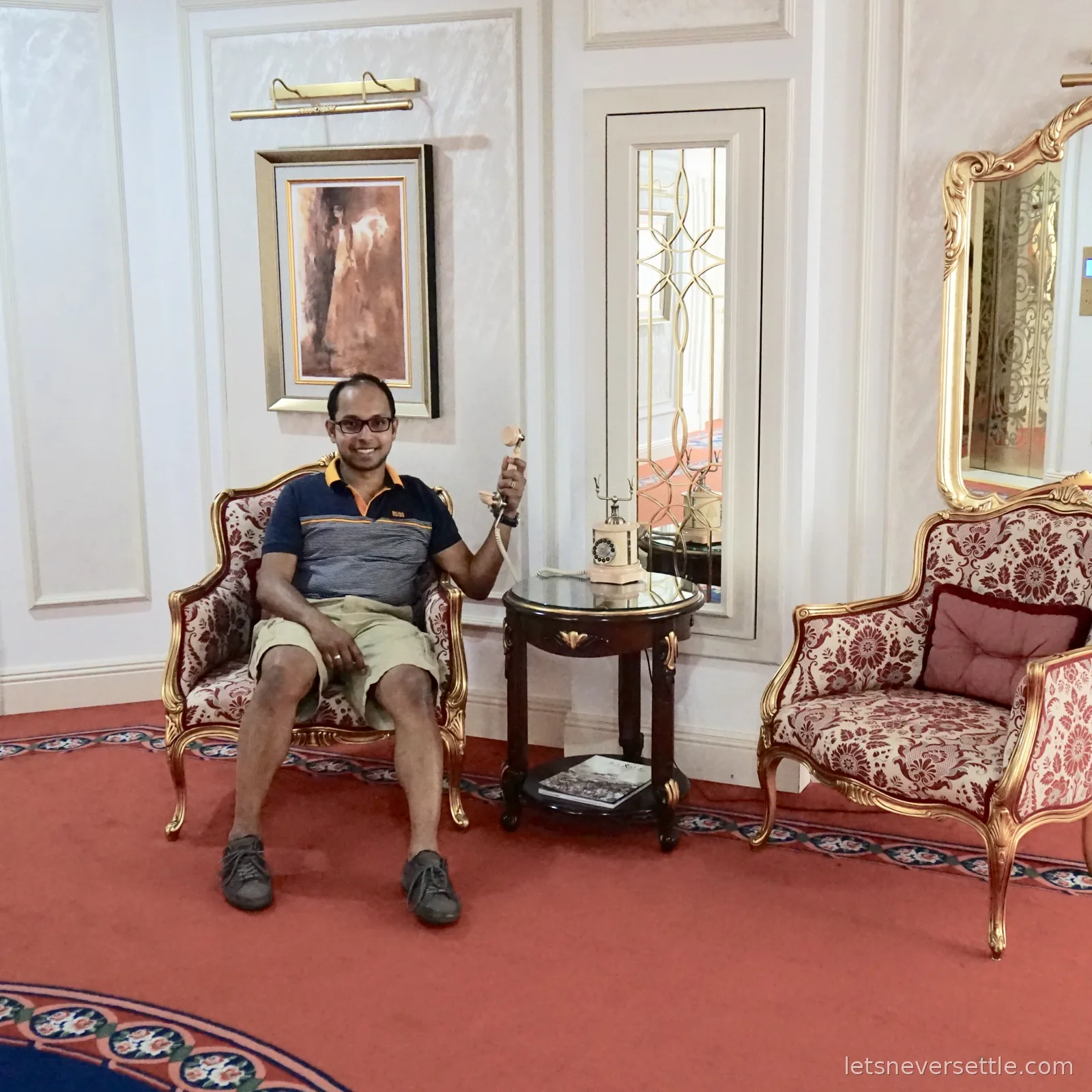 Dileep sitting in a gilded armchair holding the receiver of a vintage rotary telephone, mirrors and patterned armchairs around him