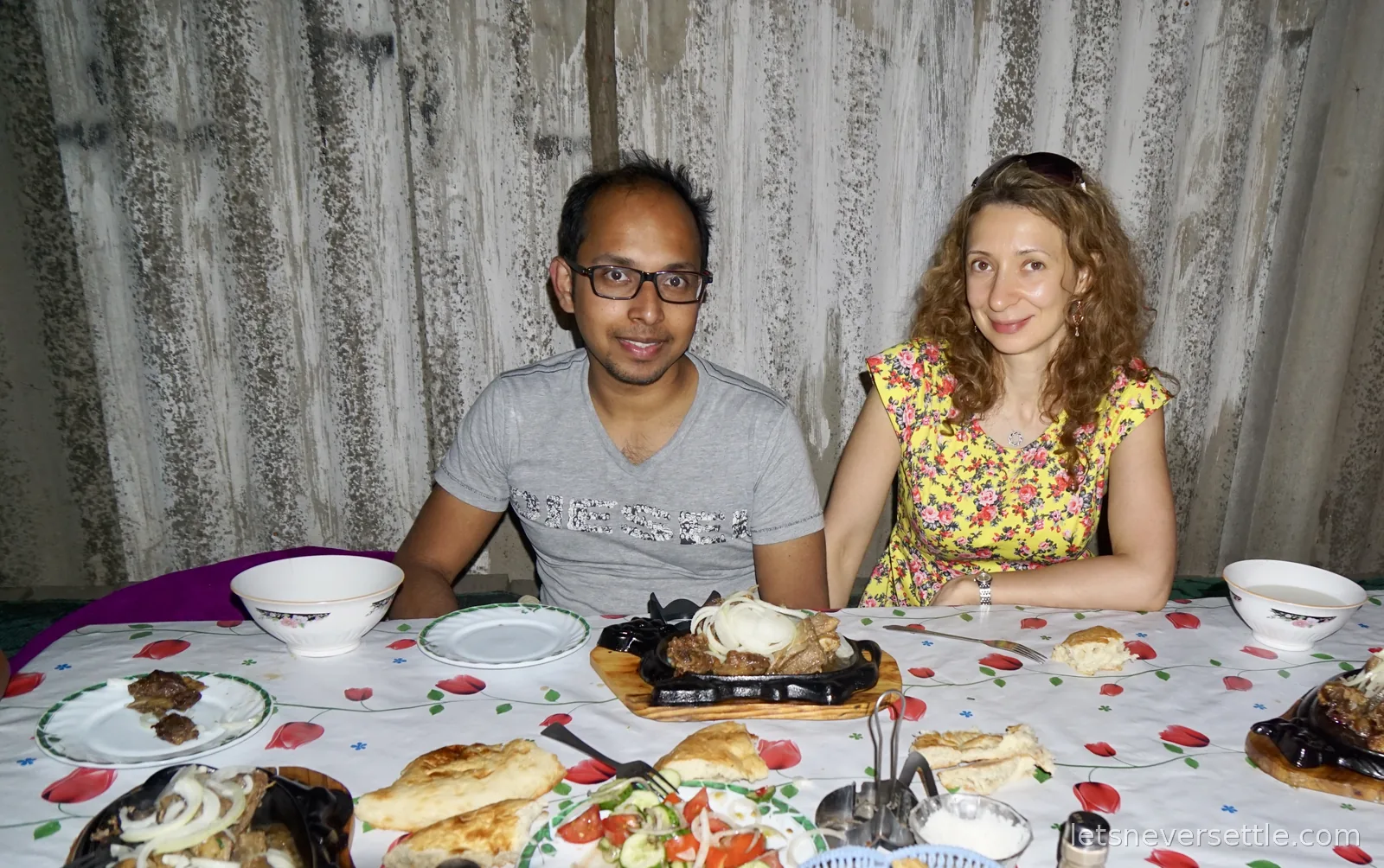 Inga and Dileep at a table with sizzling cast iron plates of meat topped with onion rings, flatbread and salad between them