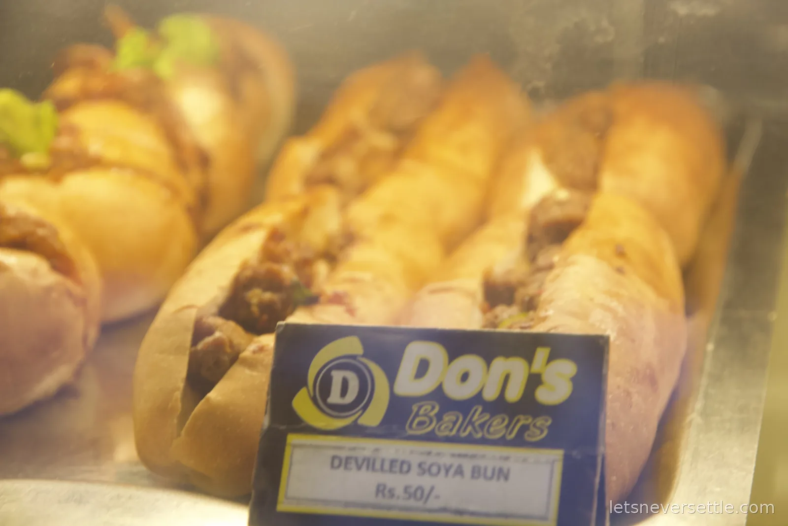 Glass bakery display case of filled buns with a Don's Bakers sign reading Devilled Soya Bun, Rs. 50