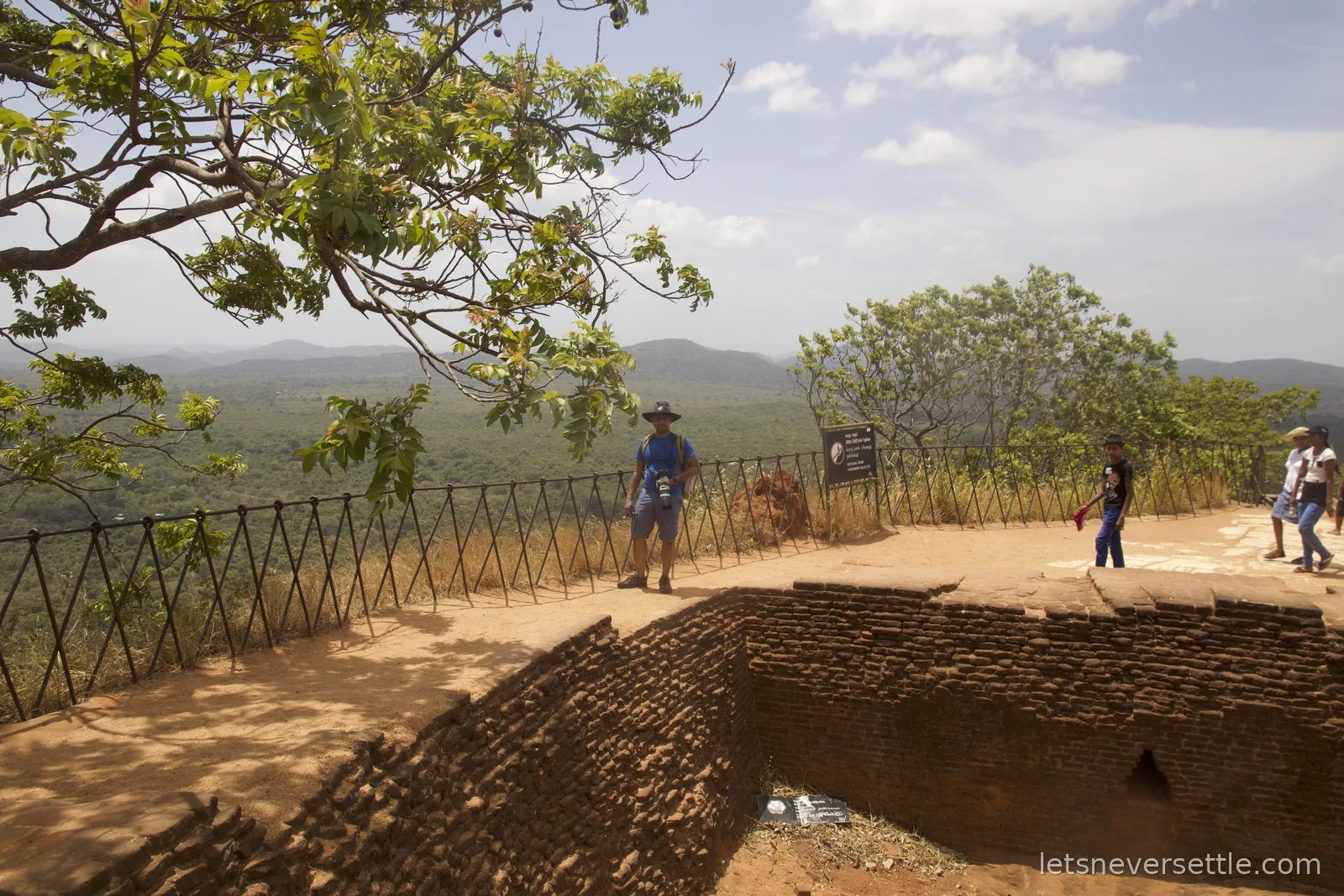 Dileep standing by the railing at the summit edge of Sigiriya with brick foundations and the jungle plain far below