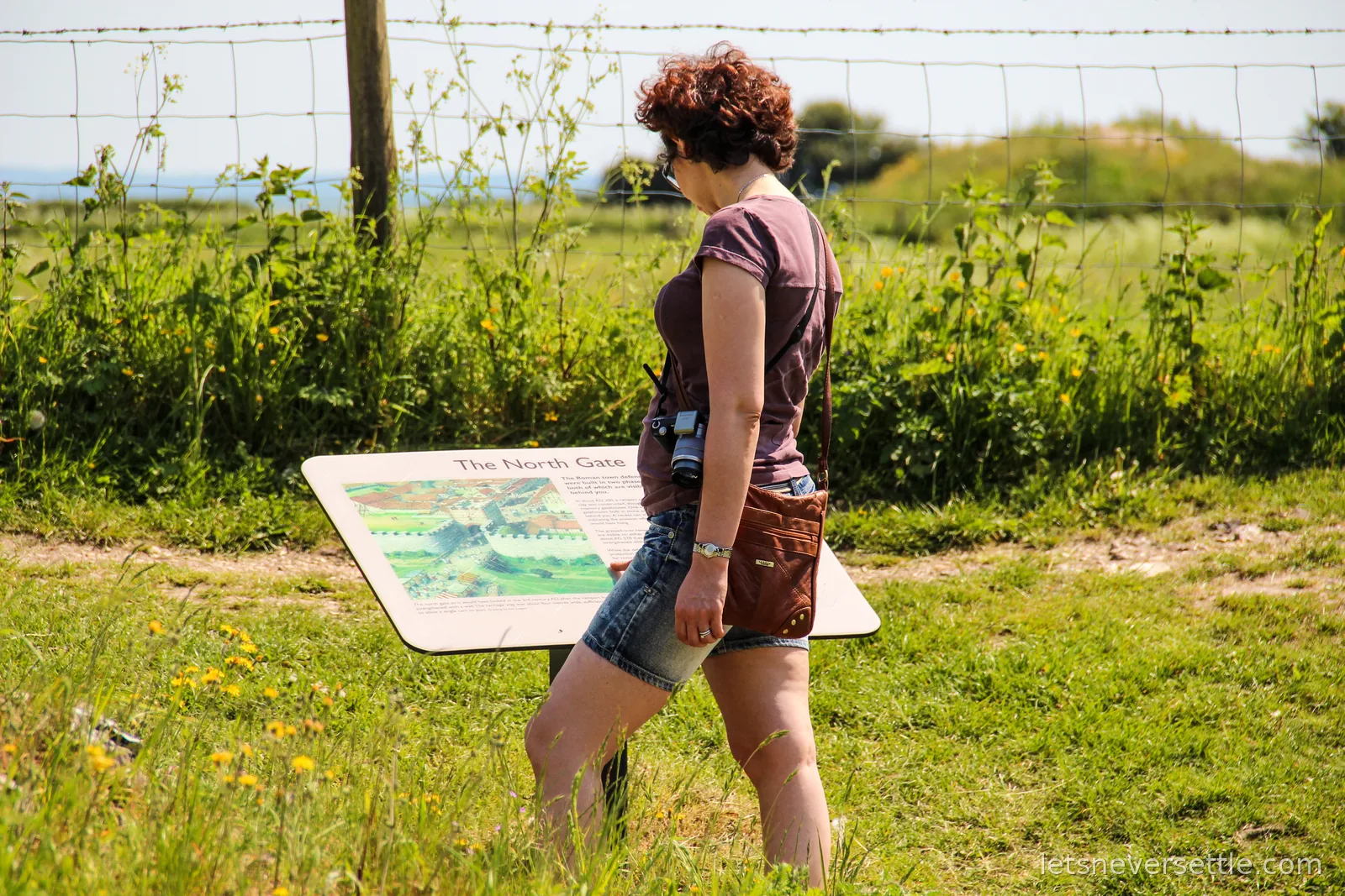 Inga with a camera over her shoulder reading an interpretation board titled The North Gate, with grass banks and fields behind