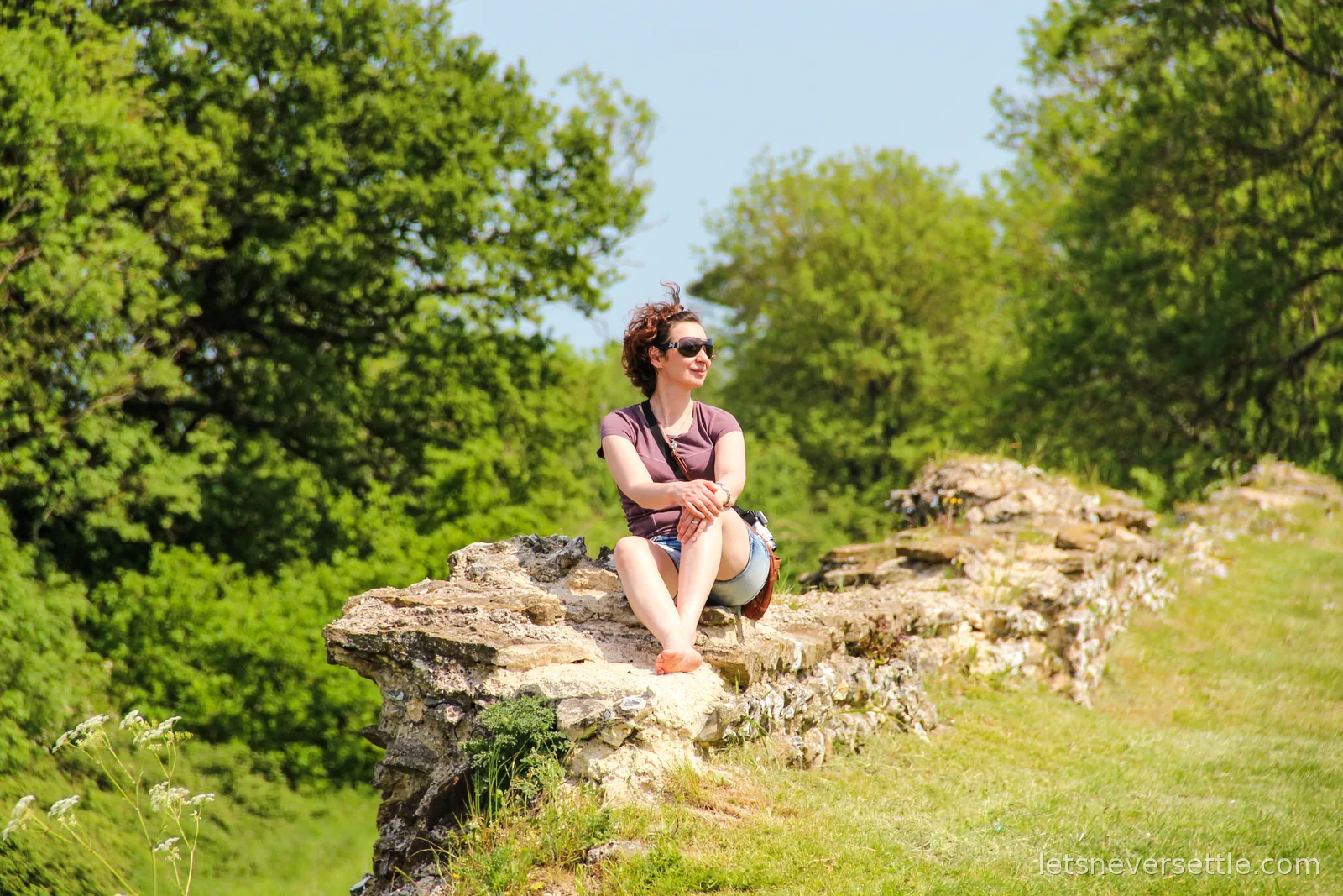 Inga in sunglasses sitting barefoot on the flint and stone remains of the Roman town wall at Silchester, green trees behind