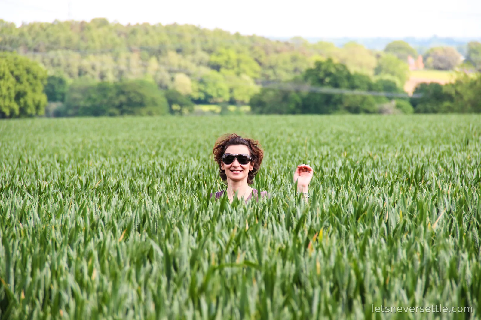 Inga in sunglasses waving from the middle of a tall green wheat field, woods and hedgerows on the horizon