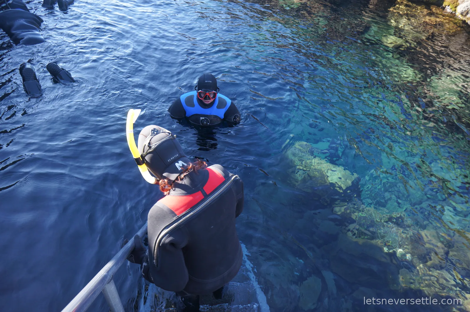 Two snorkellers in dry suits floating beside the entry steps at Silfra, with submerged rocks visible through green-blue water