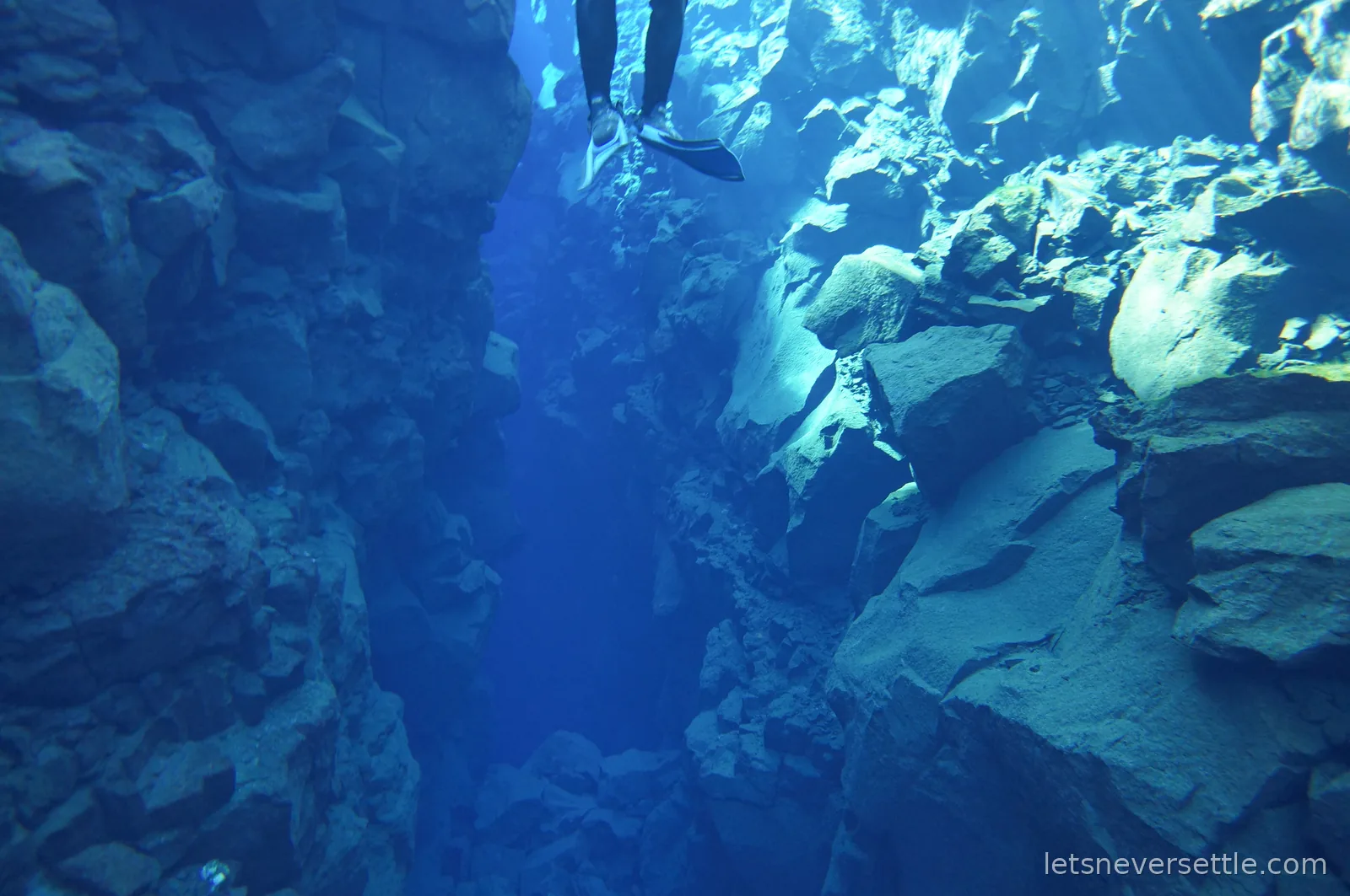 Underwater shot looking along the narrow Silfra crack as it drops into deep blue, one snorkeller finning above