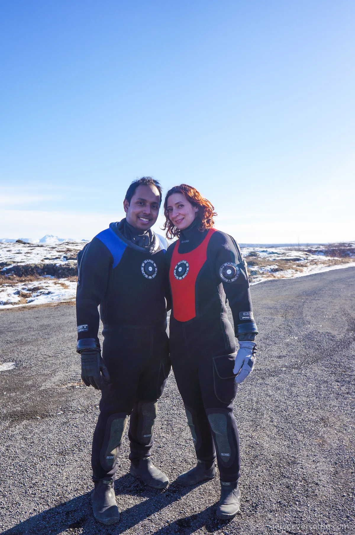 Inga and Dileep standing on a gravel road at Thingvellir in black dry suits, snow patches and blue sky behind them