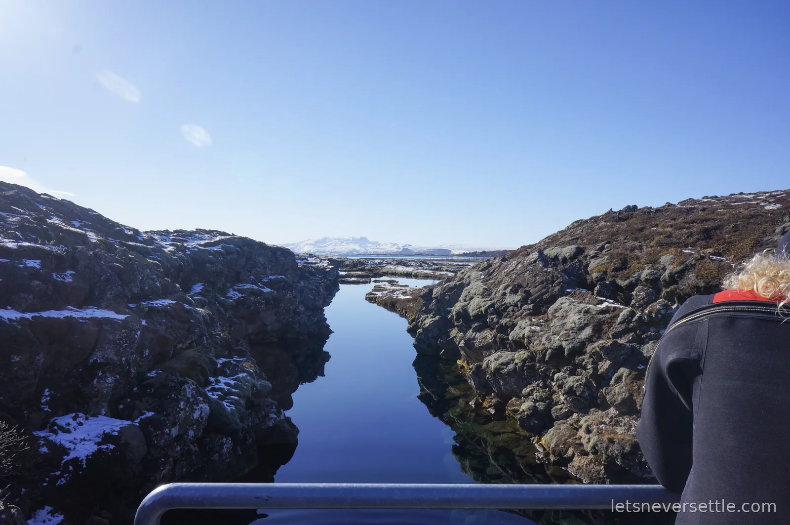 The still, dark water of the Silfra fissure running between snow-dusted rocky banks, with snow-covered mountains on the horizon