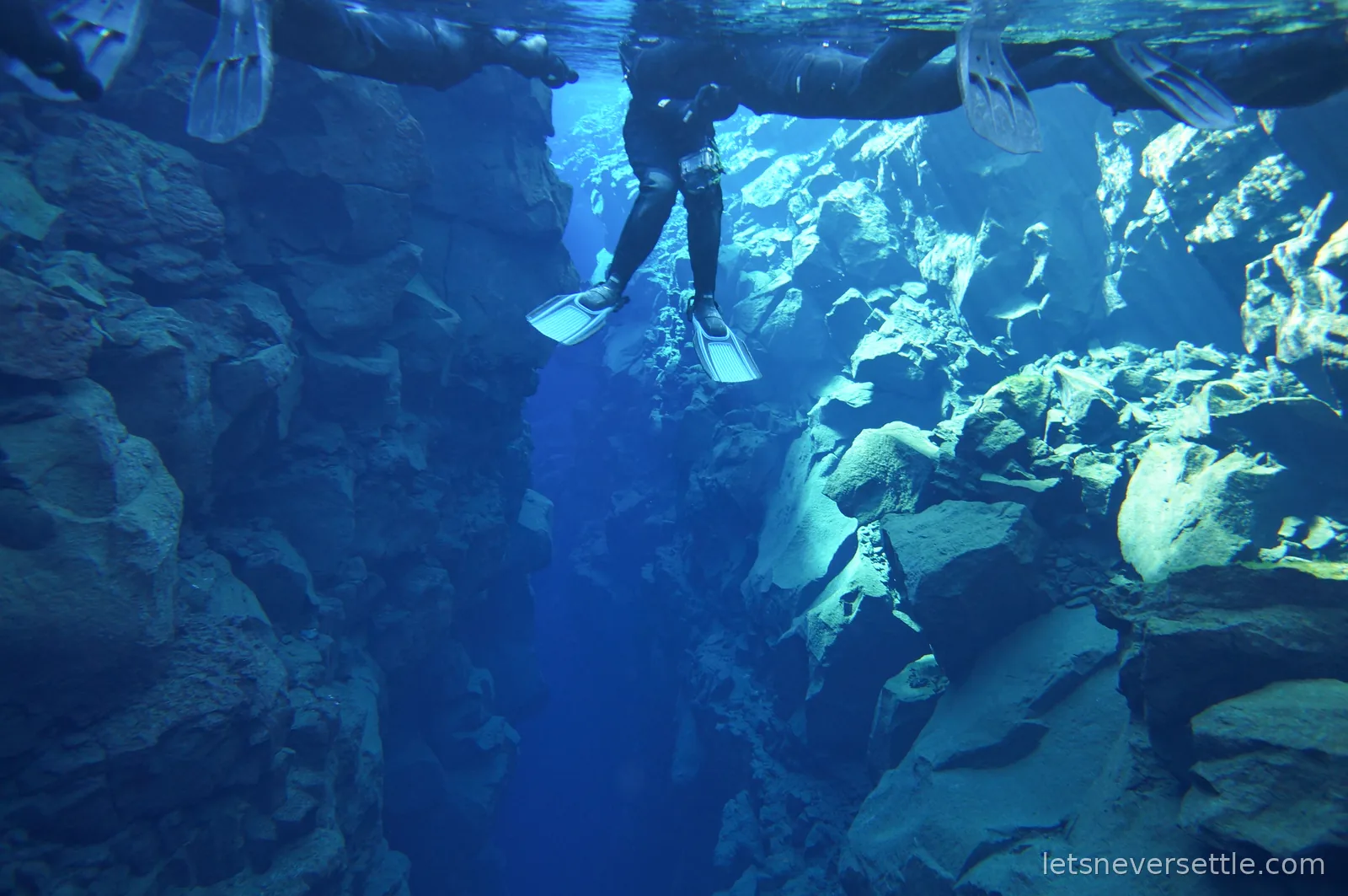 Underwater view of the Silfra fissure, rock walls on both sides with snorkellers silhouetted in fins at the surface above