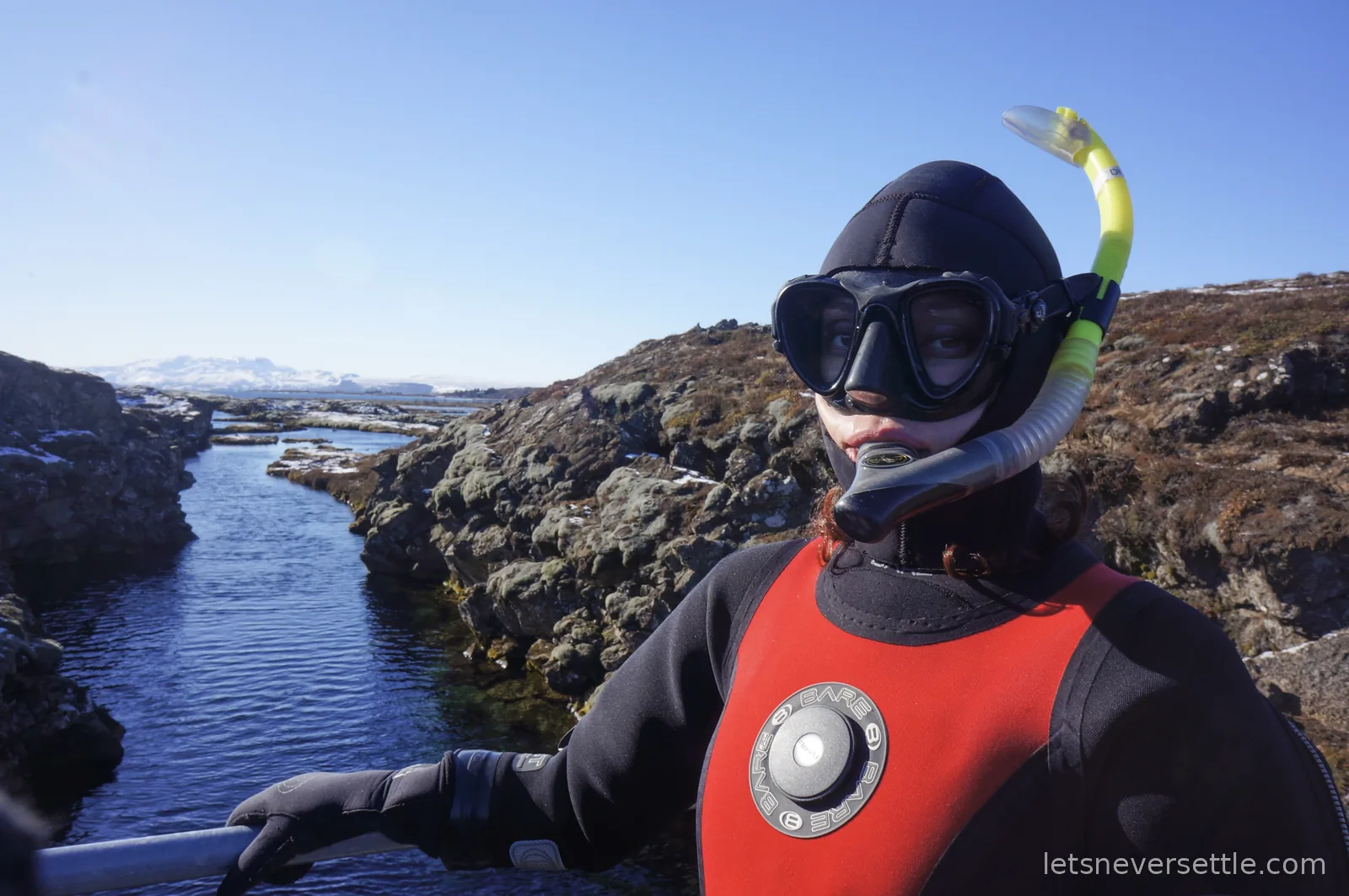 Inga in a red and black dry suit with hood, mask and yellow snorkel, holding the handrail above the Silfra fissure