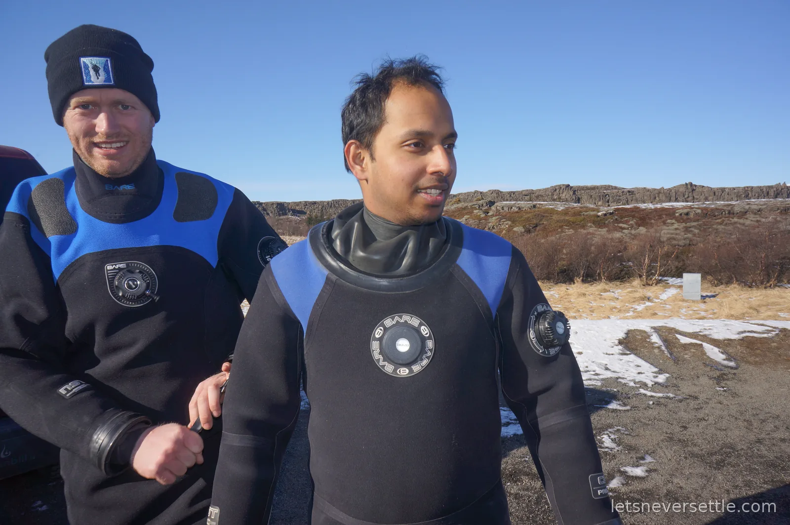 Dileep and another snorkeller in dry suits by the vehicles, with the low rocky cliffs of Thingvellir behind