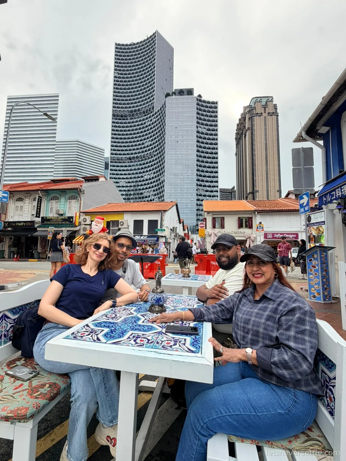 The four of us around a white table with blue Iznik-style tiles at a pavement cafe on Arab Street, shophouses, skyscrapers and a rooftop Santa behind