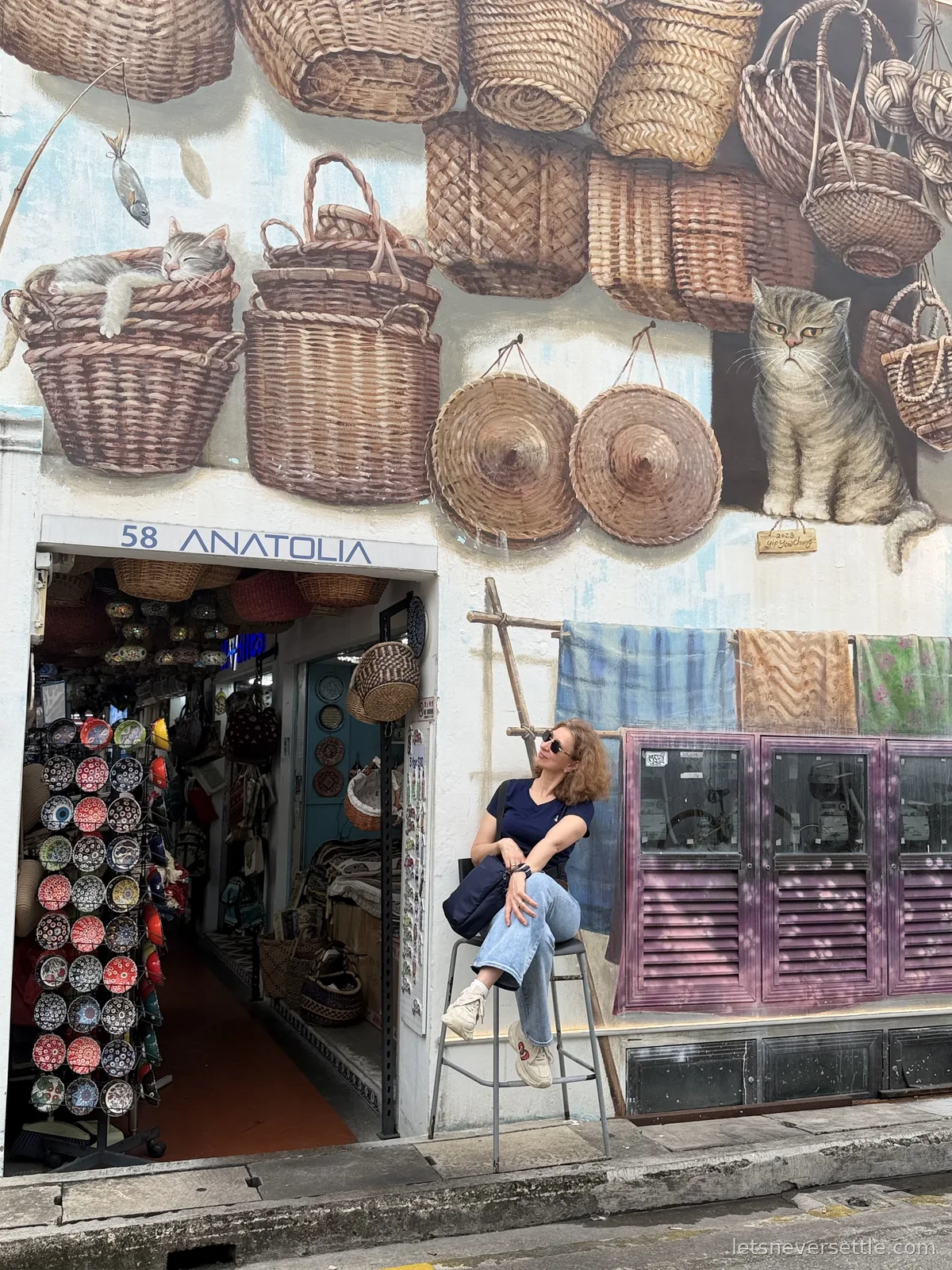 Inga sitting on a tall stool outside a Kampong Glam shop numbered 58, under a painted mural of woven baskets and two cats, with hats and rugs hung on the wall