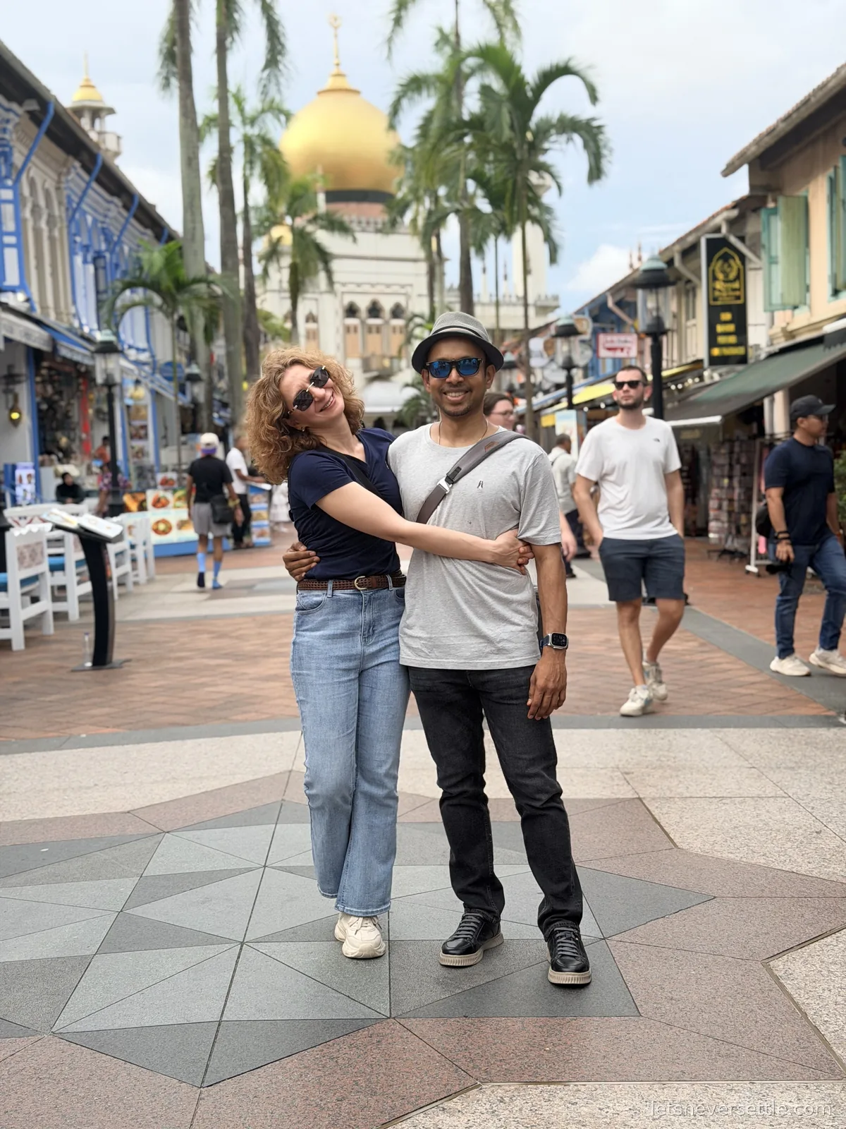 Inga and Dileep hugging on the palm-lined pedestrian street in Kampong Glam, the golden dome of the Sultan Mosque rising behind