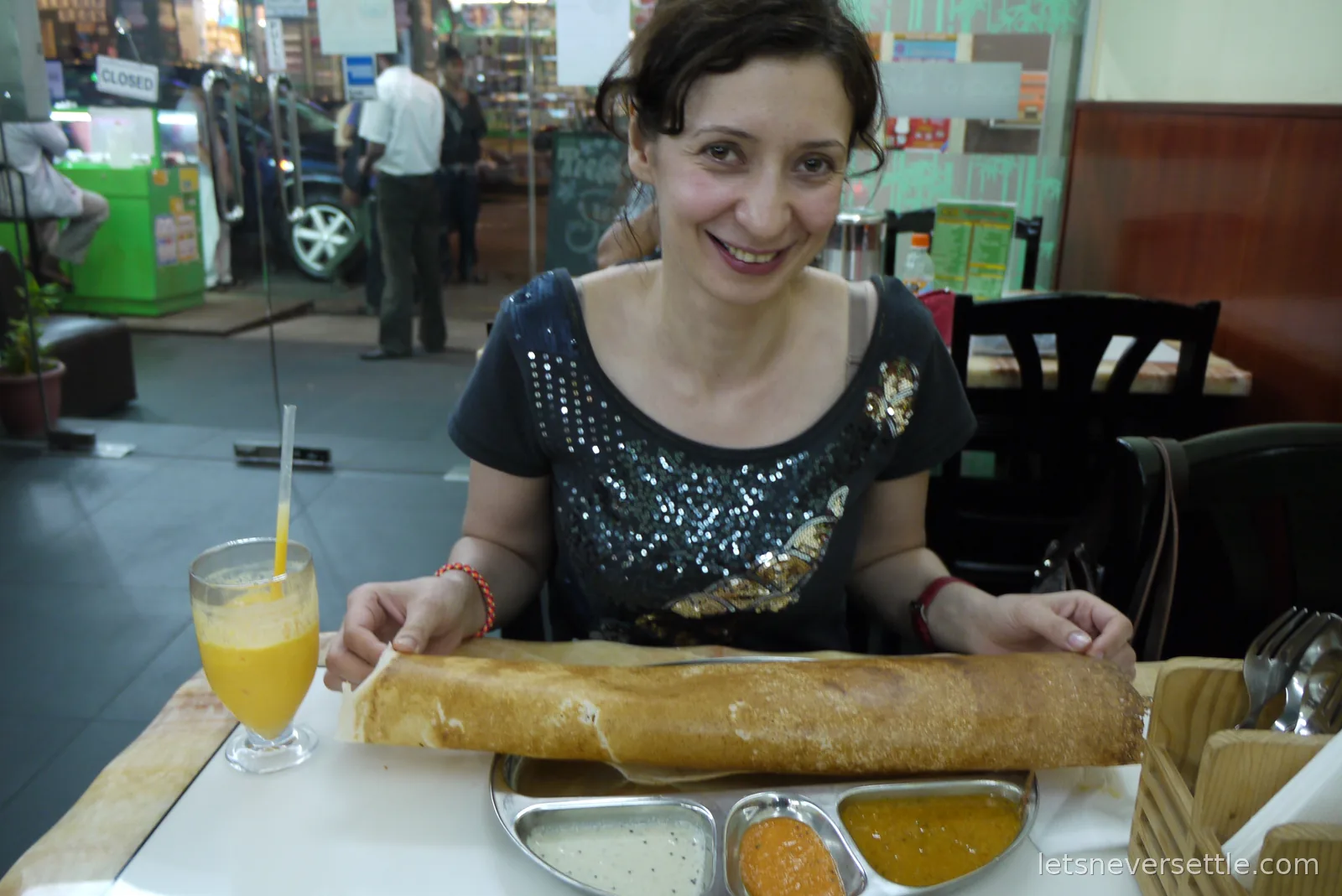 Inga smiling behind a paper dosa longer than the tray of chutneys under it, with a mango lassi on the table