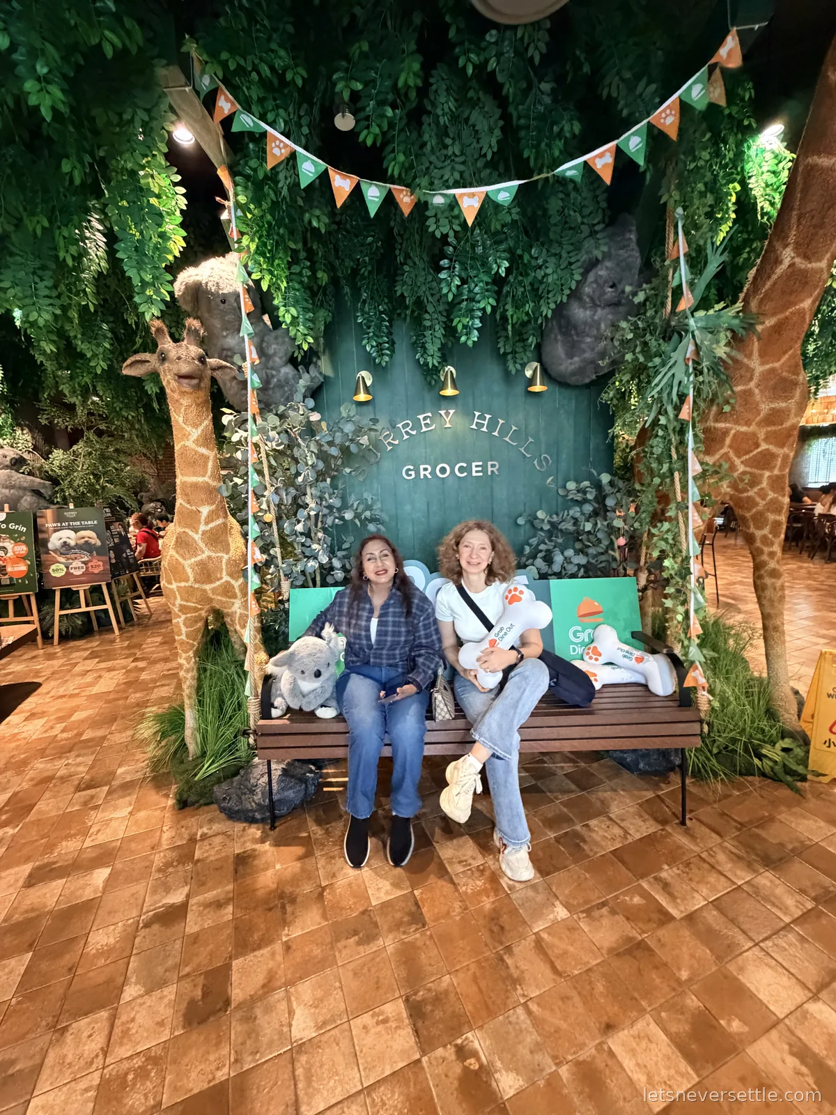 Inga and a family member sitting on a bench between two life-size giraffe models under bunting and hanging greenery at the Surrey Hills Grocer in VivoCity