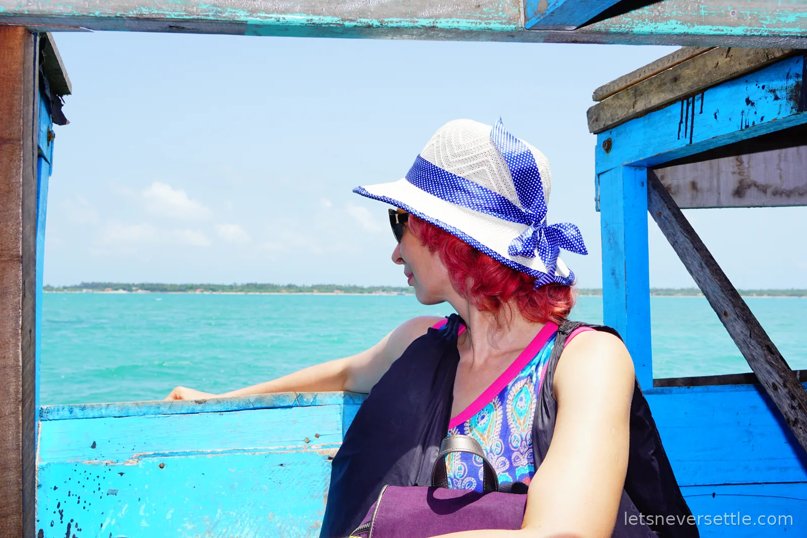 Inga in a white and blue sun hat and sunglasses sitting under the wooden roof of a bright blue open boat, looking out at flat turquoise sea with a low palm-covered shore on the horizon