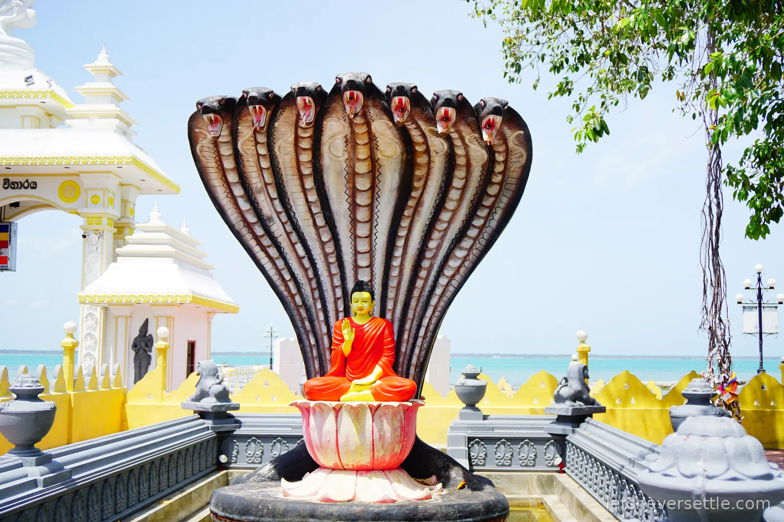 A seated Buddha in a red robe on a pink lotus with one hand raised, sheltered by a huge seven-headed cobra canopy, with white temple buildings on the left and turquoise sea behind