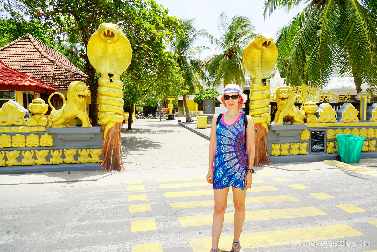 Inga in a blue patterned dress and sun hat standing on the road between two towering golden cobra statues with flared hoods that serve as the gateposts of a Buddhist temple