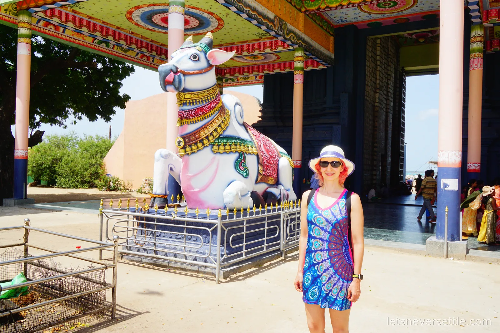 Inga standing in front of the Nandi bull statue under a canopy painted with red, green and gold roundels, with pilgrims sitting in the shade of the temple doorway behind