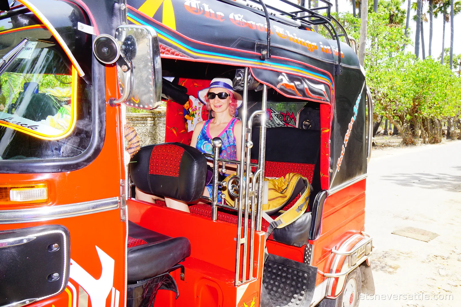 Inga sitting in the back of a red tuk-tuk decorated with airbrushed lettering and chrome bars, parked on a sandy road lined with palmyra palms