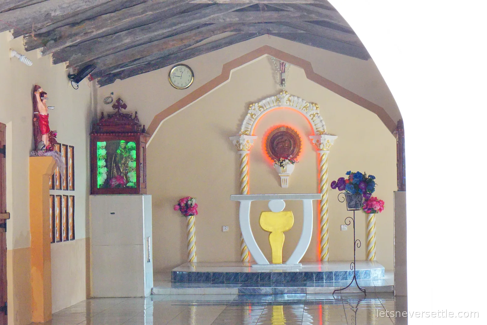 The tiled interior of a small Catholic church with rough wooden roof beams, a figure of Christ on a plinth, a wooden shrine cabinet lit green, and a white altar niche with a glowing red halo