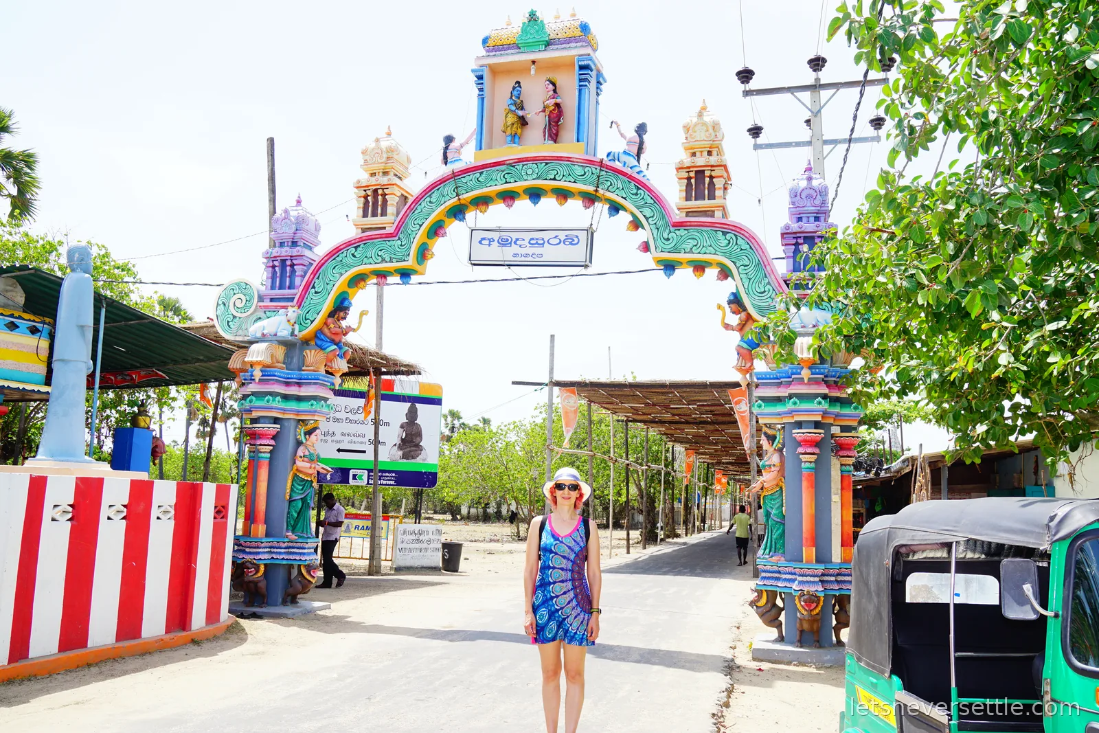 Inga standing on a sandy road beneath a tall painted Hindu archway covered in turquoise, pink and gold figures, with a green road sign and a green tuk-tuk to one side