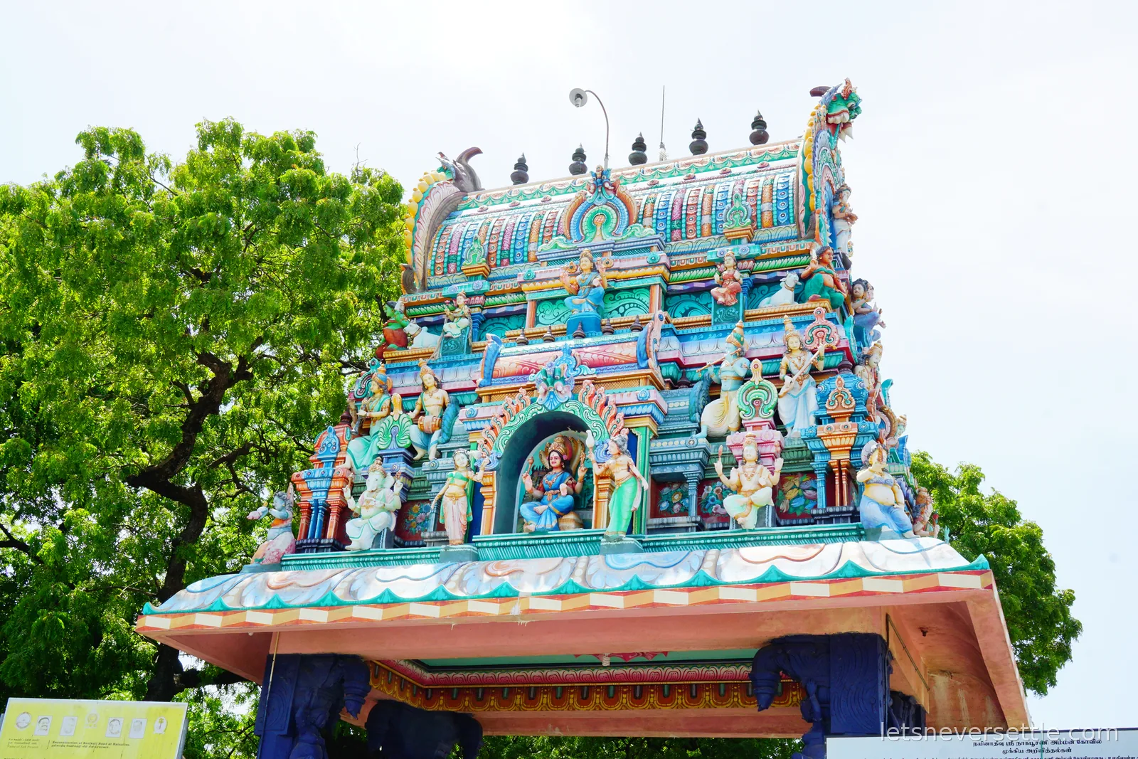 A tall temple gopuram tower covered in hundreds of brightly painted sculpted figures in turquoise, pink, gold and green, seen from below against a white sky