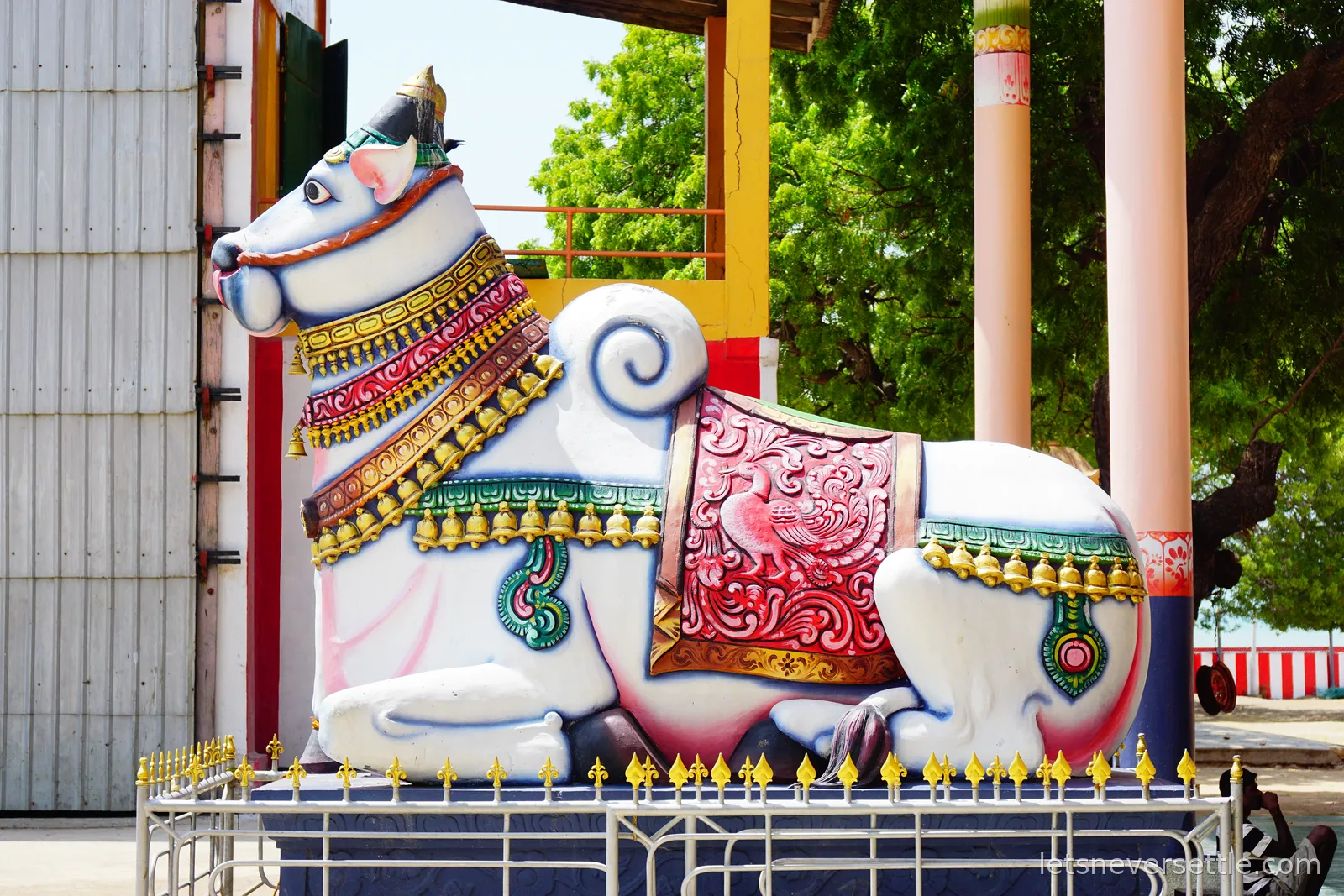A large white and pink statue of Nandi the bull lying down, wearing a red embroidered saddle cloth, gold bells and a jewelled collar, behind a low fence of gold spear tips