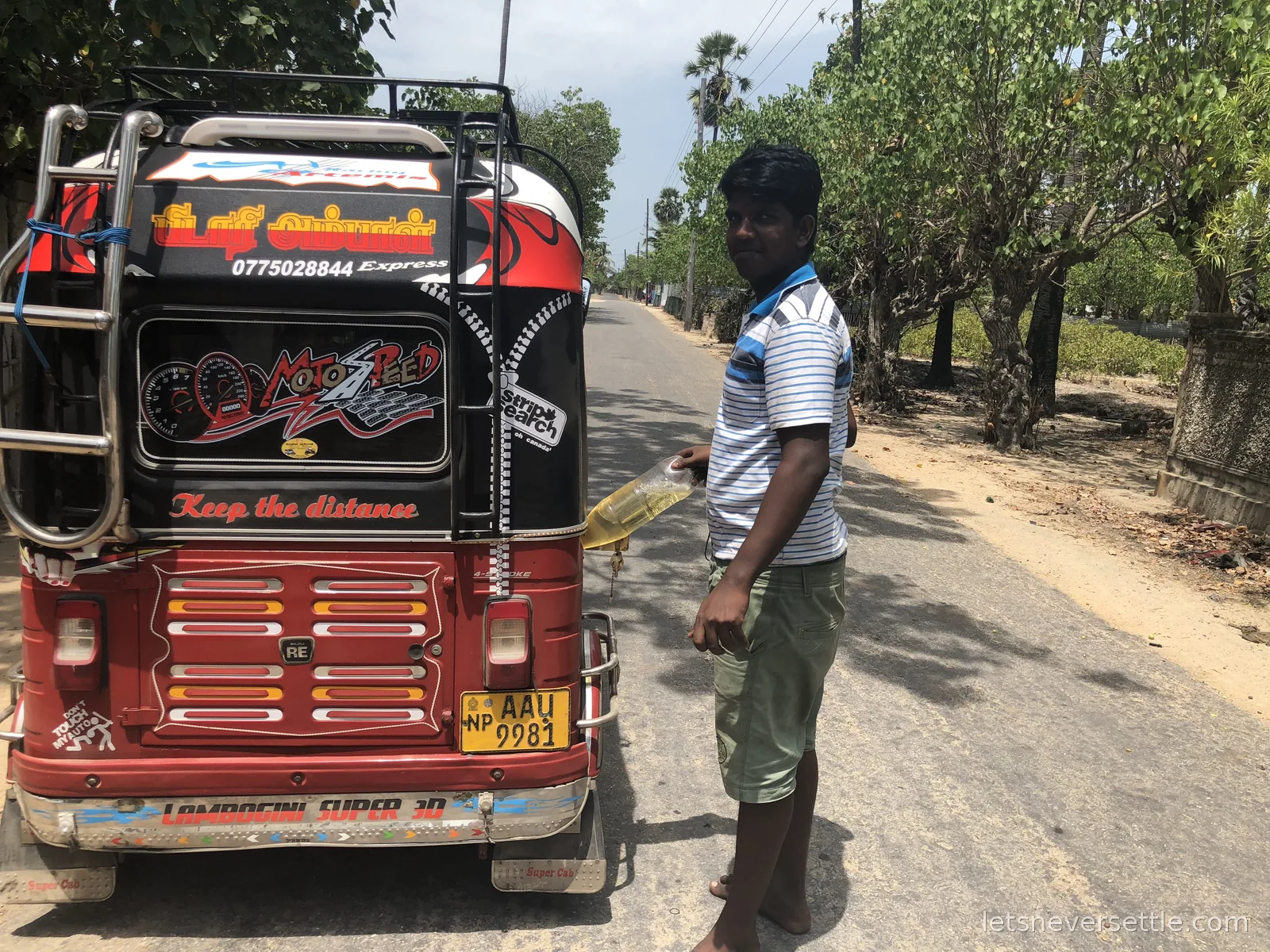 A young man in a striped polo shirt standing barefoot beside a red and black tuk-tuk on a quiet island road, holding a clear plastic bottle of yellow petrol