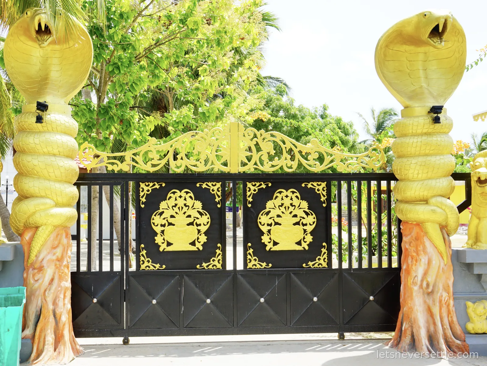 A close view of the black and gold temple gate flanked by two giant golden cobra statues coiled around painted tree trunks, with gilded scrollwork above the railings