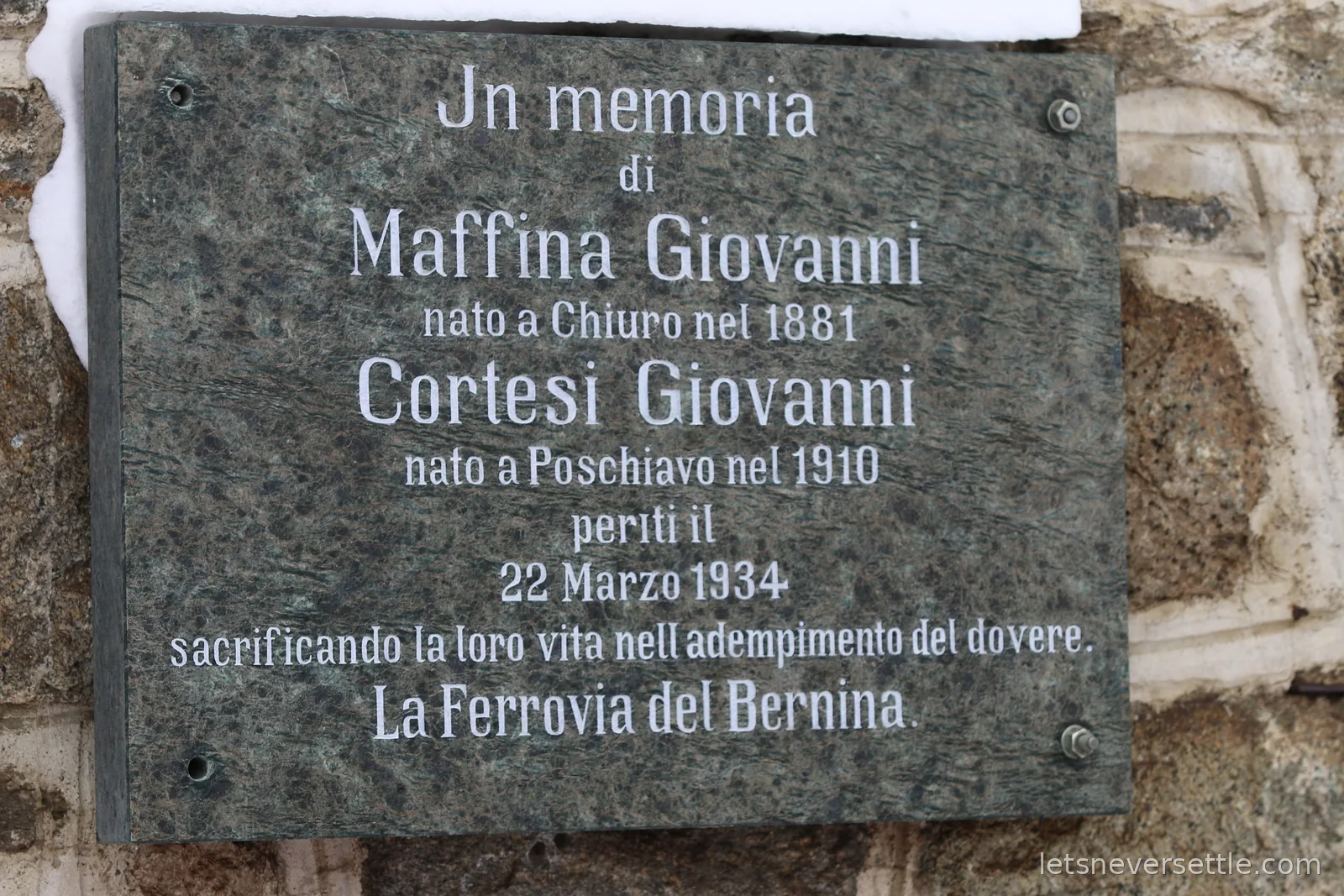 A dark stone plaque set into a rough wall, in memory of Giovanni Maffina and Giovanni Cortesi, who died on 22 March 1934, placed by the Bernina Railway