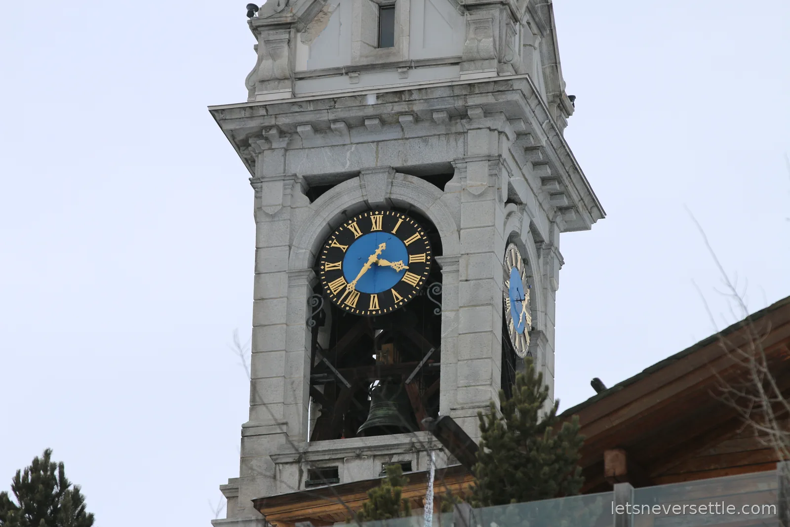 A pale stone bell tower with a blue and gold clock face and a bronze bell visible in the open belfry