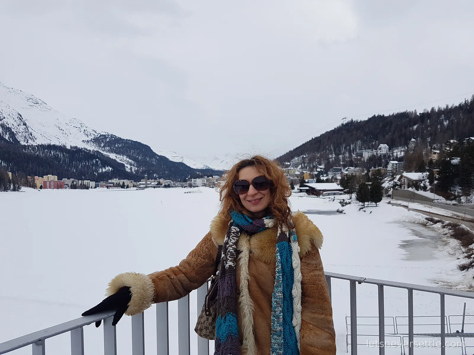Inga leaning on a white railing in a shearling coat and blue scarf, with a completely frozen snow covered lake behind her and the town on the far shore